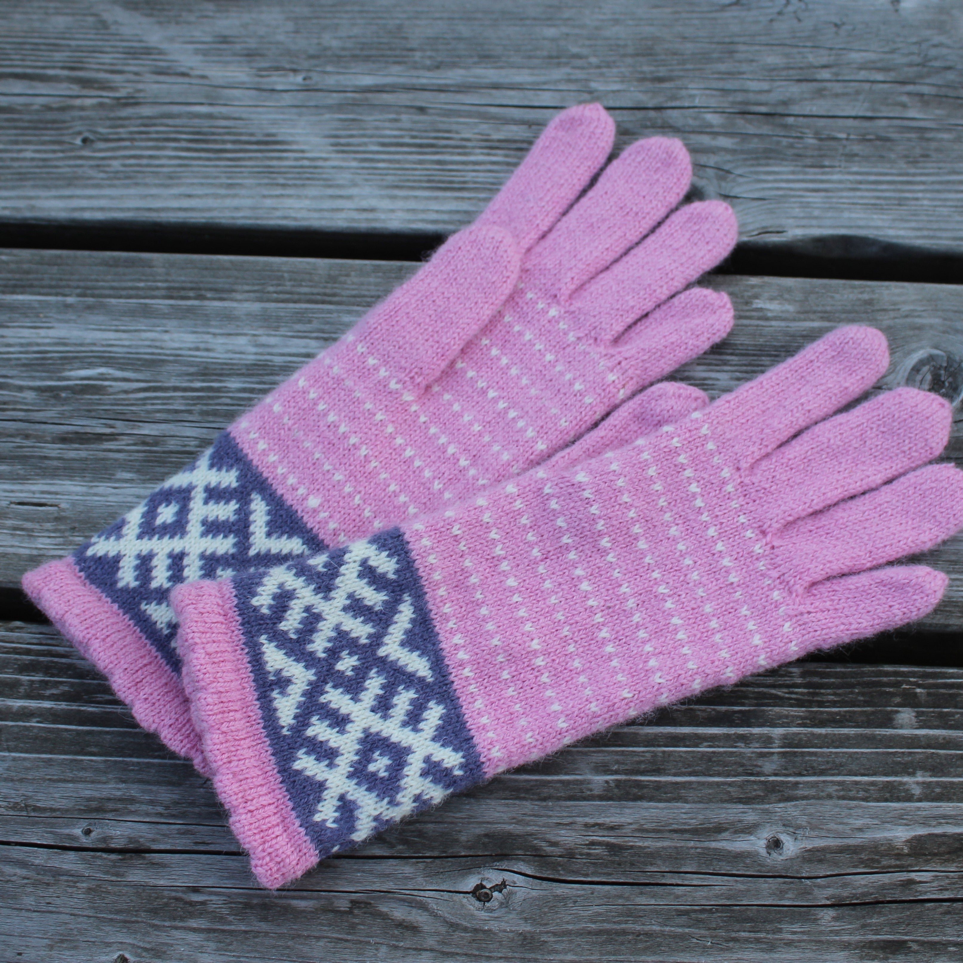 A pair of pink knit gloves with a white geometric pattern on the cuffs, laid flat on a weathered gray wooden surface.