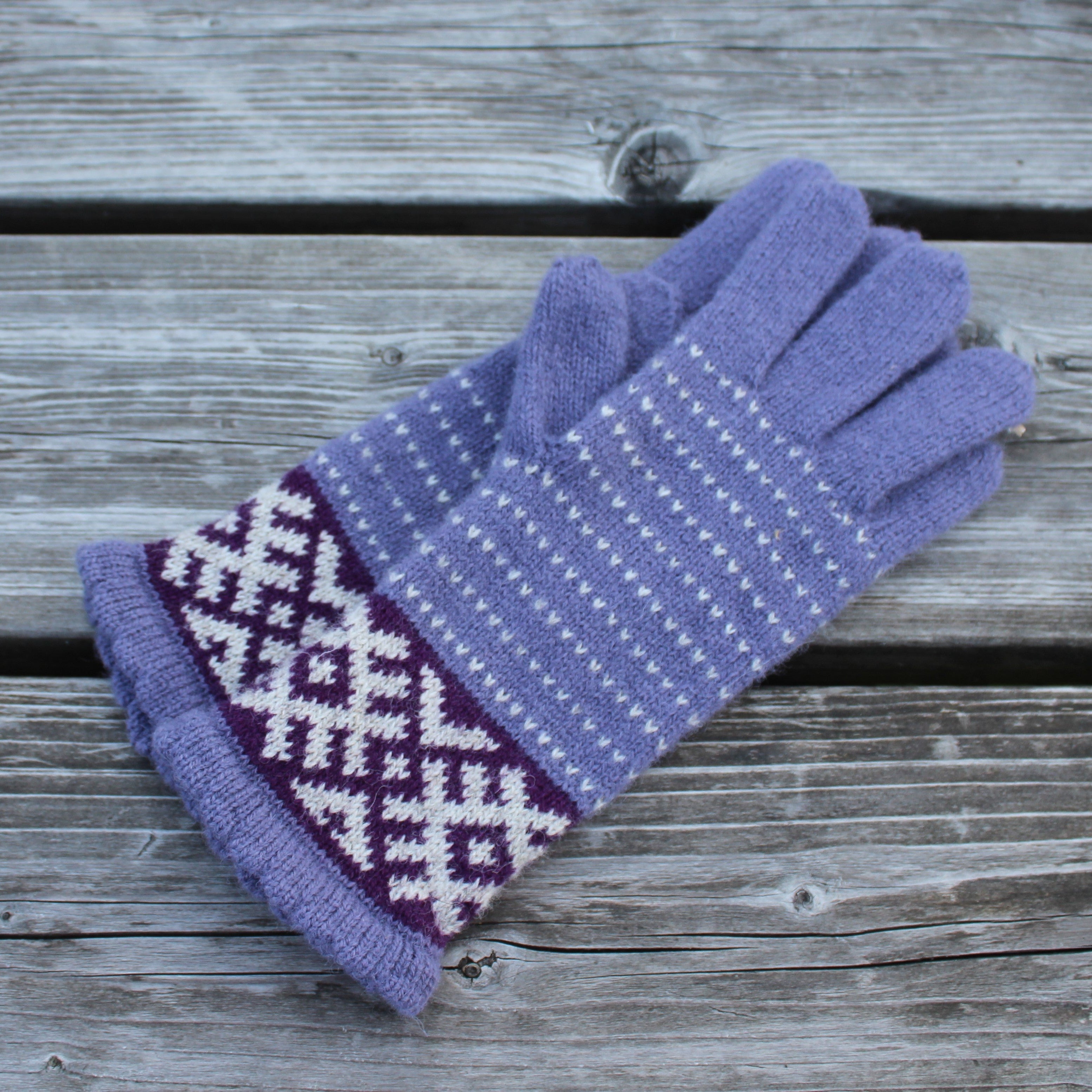 A pair of knit gloves in lavender purple with white geometric patterns and a darker purple cuff, displayed on a weathered gray wooden surface.