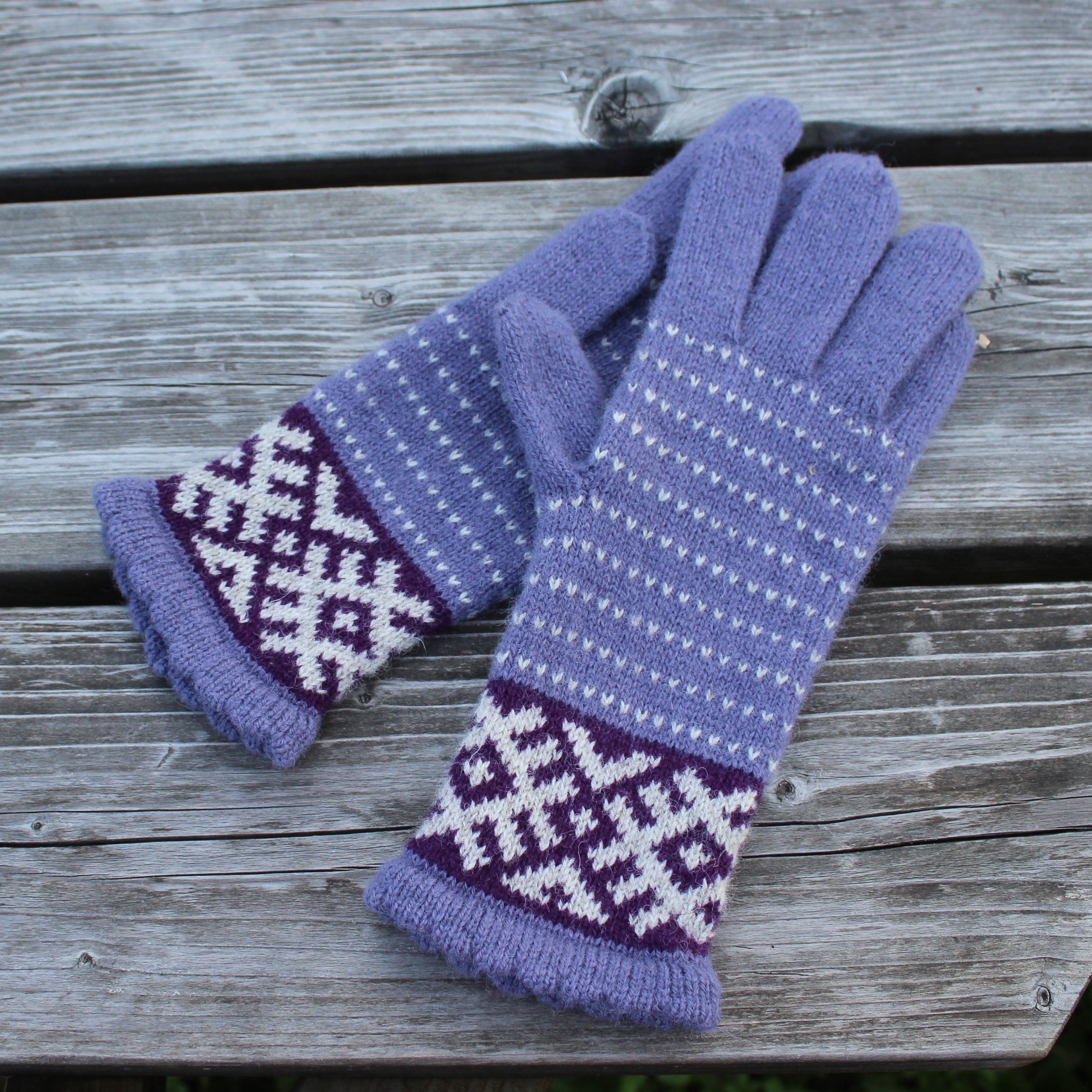 A pair of purple knit winter gloves with white and dark purple geometric patterns, displayed on a weathered gray wooden plank surface.