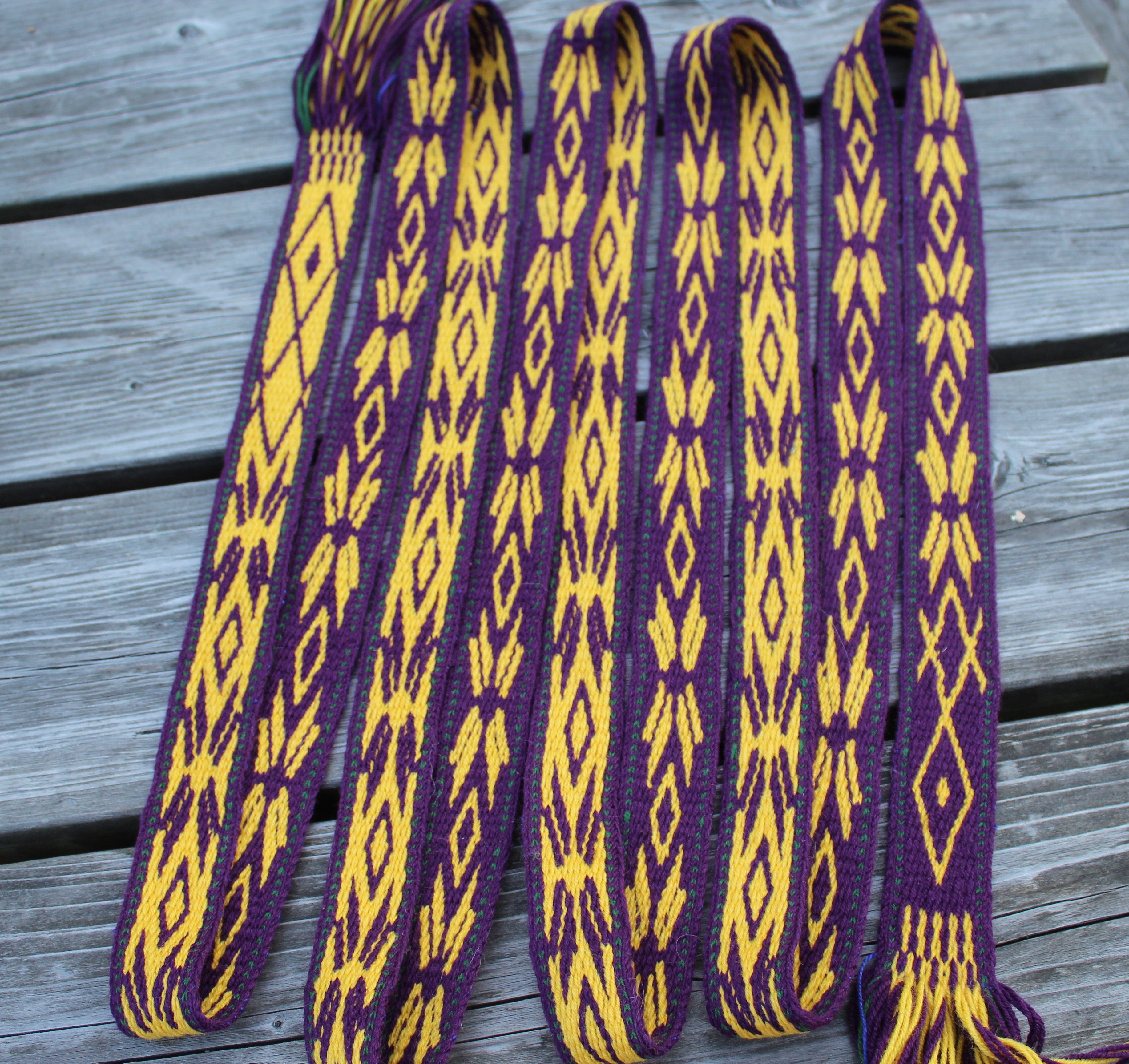 A set of purple and yellow woven strap belts with geometric diamond patterns, laid out on a gray wooden plank surface.