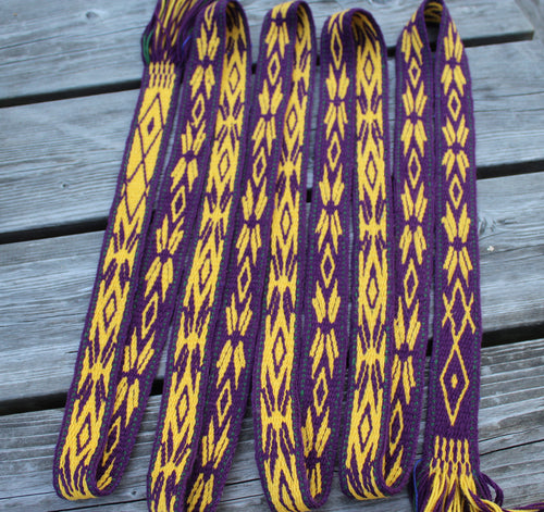 A set of purple and yellow woven strap belts with geometric diamond patterns, laid out on a gray wooden plank surface.