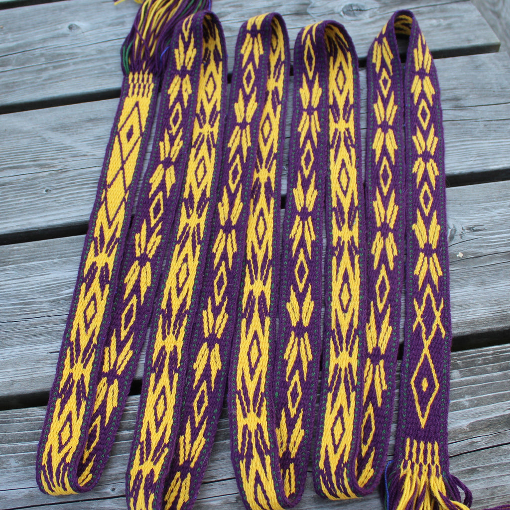 A woven Peruvian-style shoulder strap or belt with purple and yellow geometric patterns, laid out on a gray wooden deck.