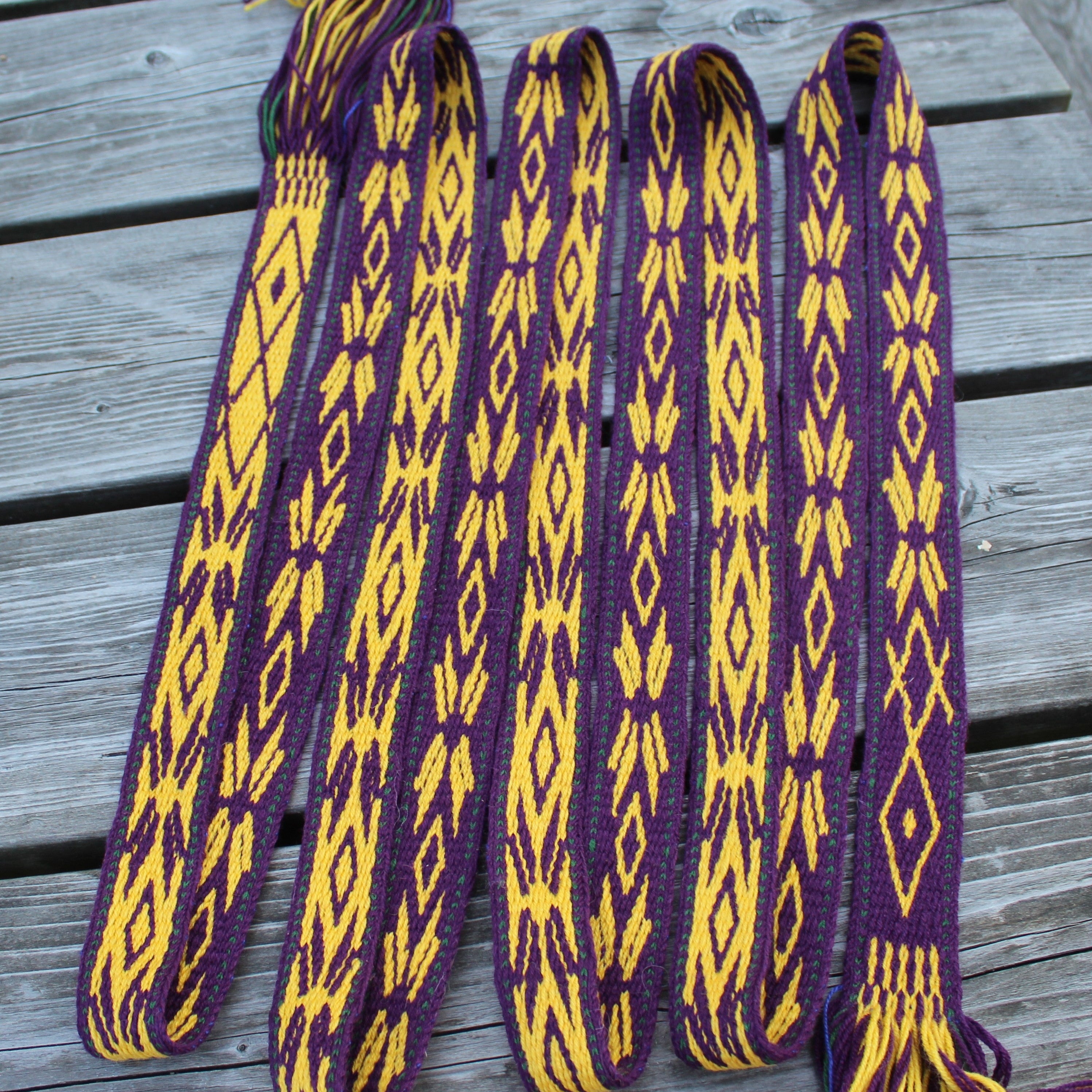 A woven Peruvian-style shoulder strap or belt with purple and yellow geometric patterns, laid out on a gray wooden deck.