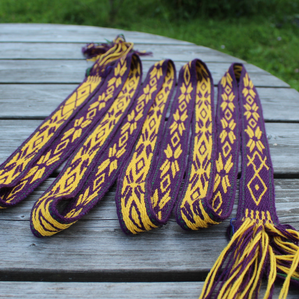 A set of five handwoven Peruvian llama wool shoulder straps (repetidores) in purple and yellow, laid out on a weathered gray wooden table with a green garden background.