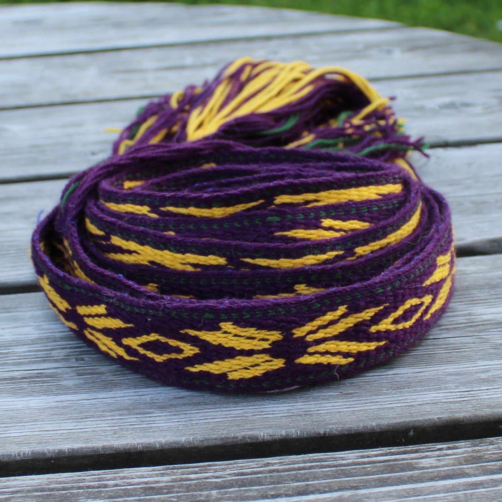 A coiled purple woven strap with yellow geometric patterns and yellow tassels, resting on a gray wooden table with green grass in the background.