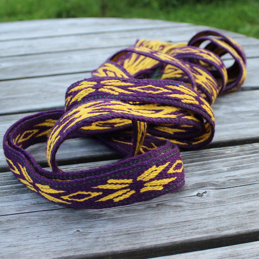 A coiled woven strap or belt with a purple and yellow geometric pattern, resting on a weathered gray wooden table with green grass in the background.