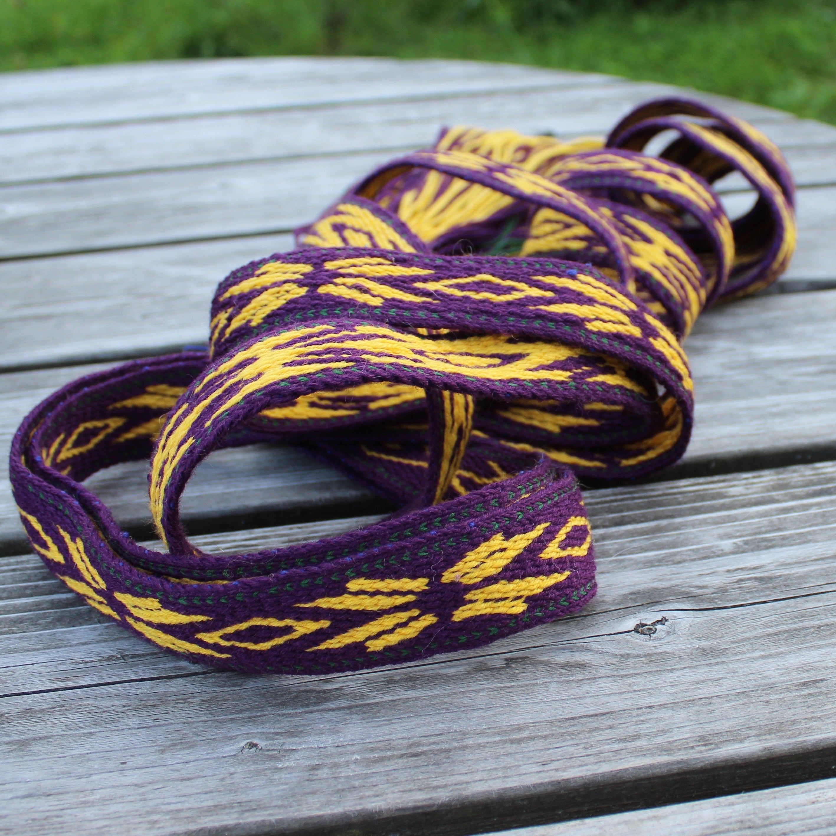 A coiled woven strap or belt with a purple and yellow geometric pattern, resting on a weathered gray wooden table with green grass in the background.