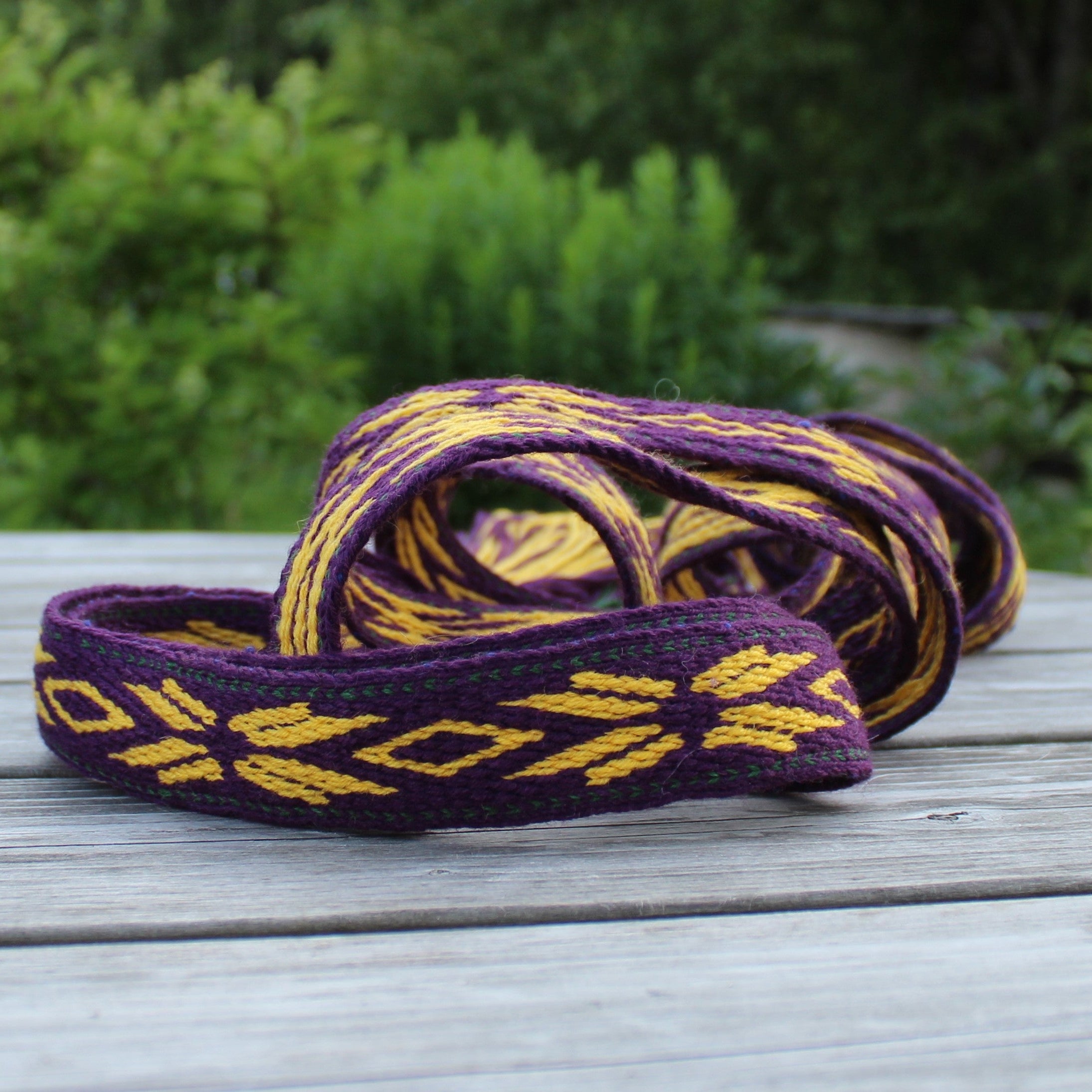 A purple and yellow woven elastic band with a geometric diamond pattern, resting on a weathered gray wooden table with a blurred green garden background.