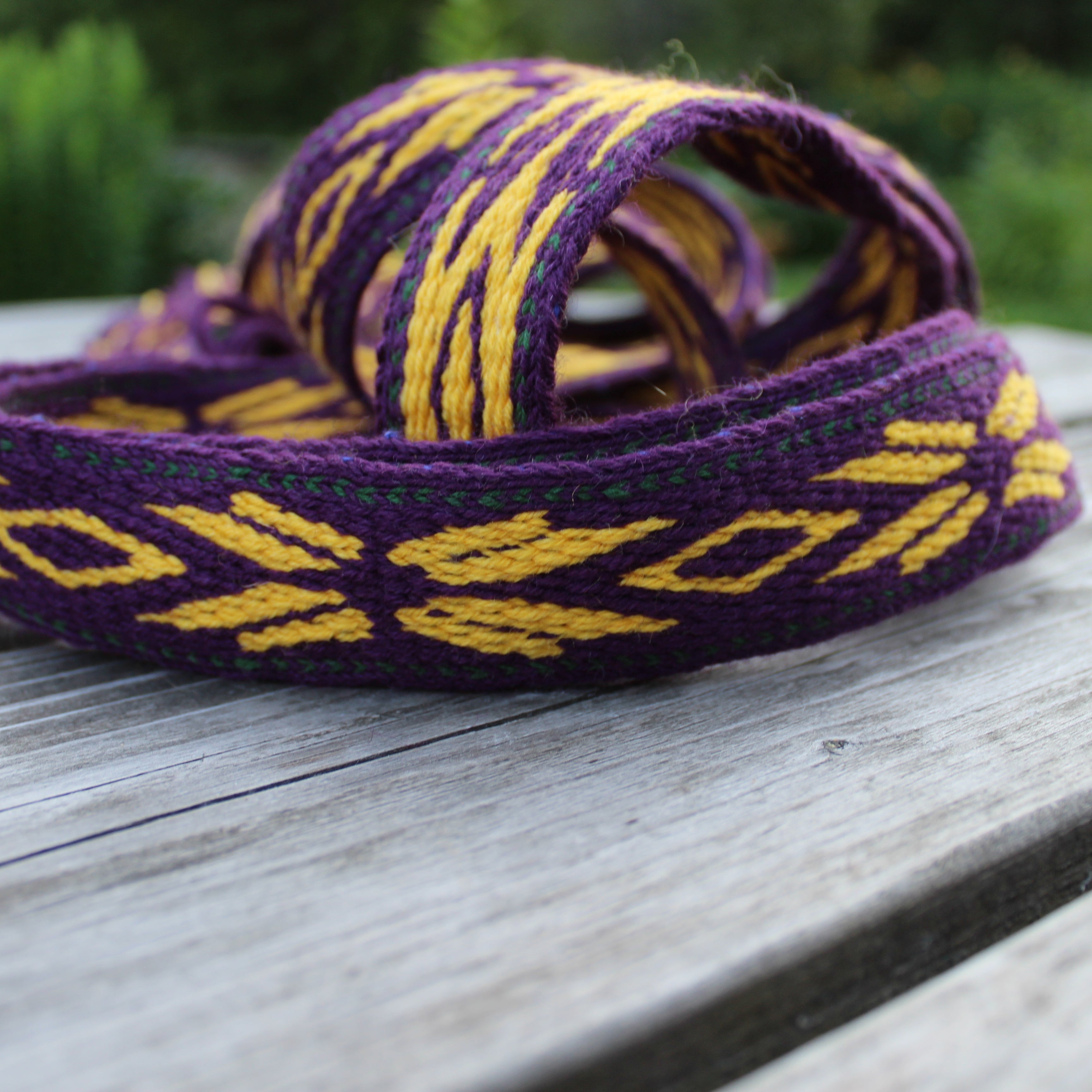 A purple and yellow woven strap or belt with geometric patterns, resting on a weathered gray wooden table with a blurred green outdoor background.