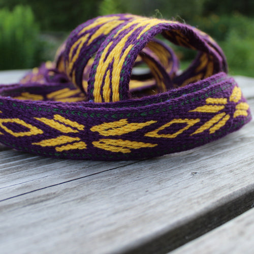 A purple and yellow woven strap or belt with geometric patterns, resting on a weathered gray wooden table with a blurred green outdoor background.