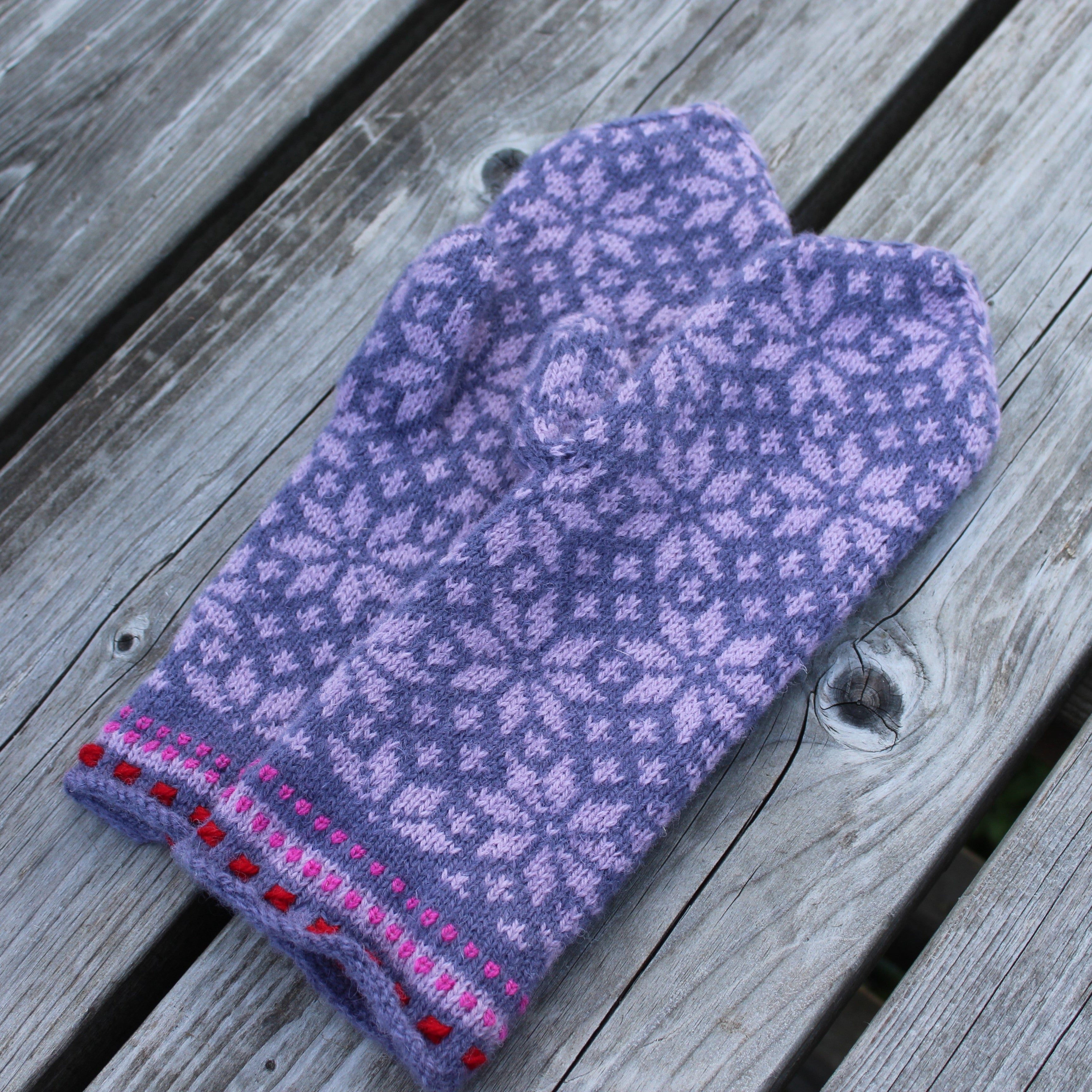 A single knit mitten in lavender and light purple with a snowflake pattern, featuring a pink and red striped cuff with red pom-poms, laid flat on weathered gray wooden planks.