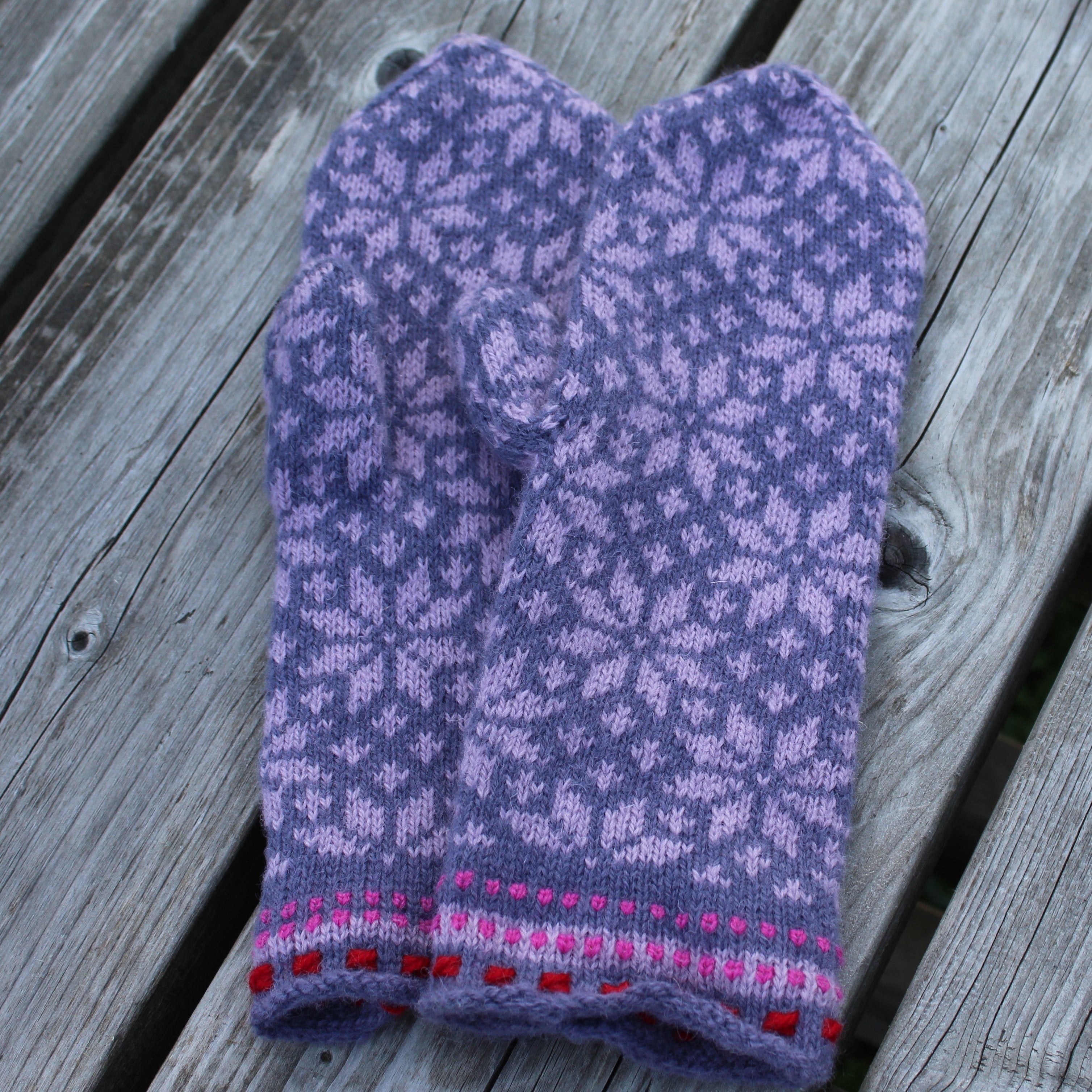 A pair of hand-knit purple and lavender mittens with a snowflake pattern and a pink and red striped cuff, laid flat on weathered gray wooden planks.