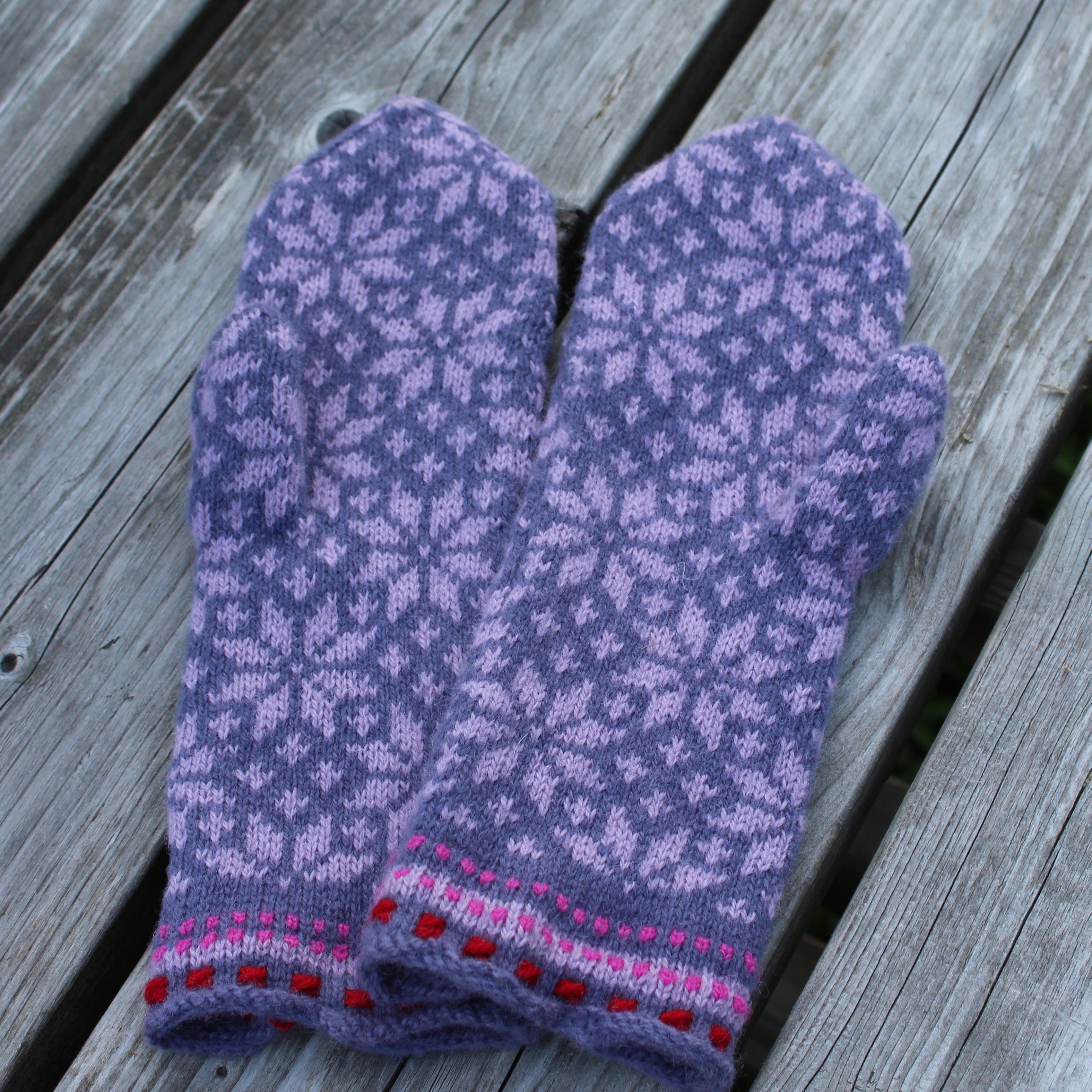 A pair of hand-knit winter mittens in a lavender-purple color with a light purple snowflake pattern and pink and red striped trim, displayed on weathered gray wooden planks.