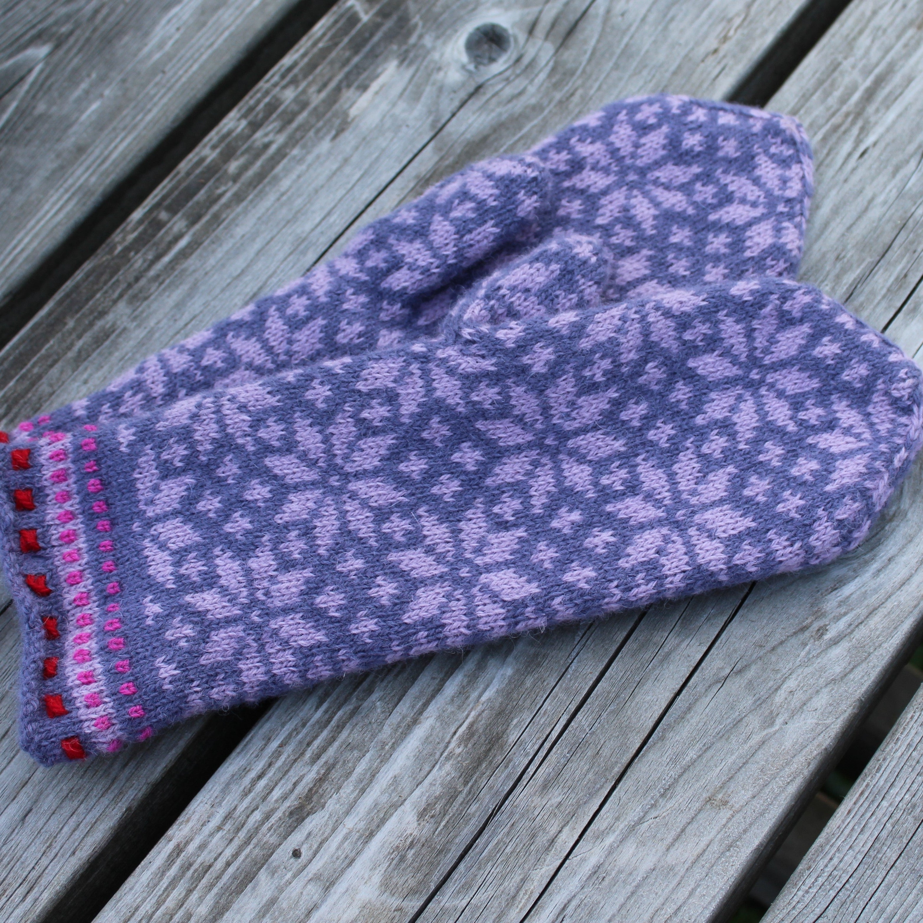 A single knit mitten in lavender purple with a light purple snowflake pattern and a pink and red striped cuff, resting on weathered gray wooden planks.