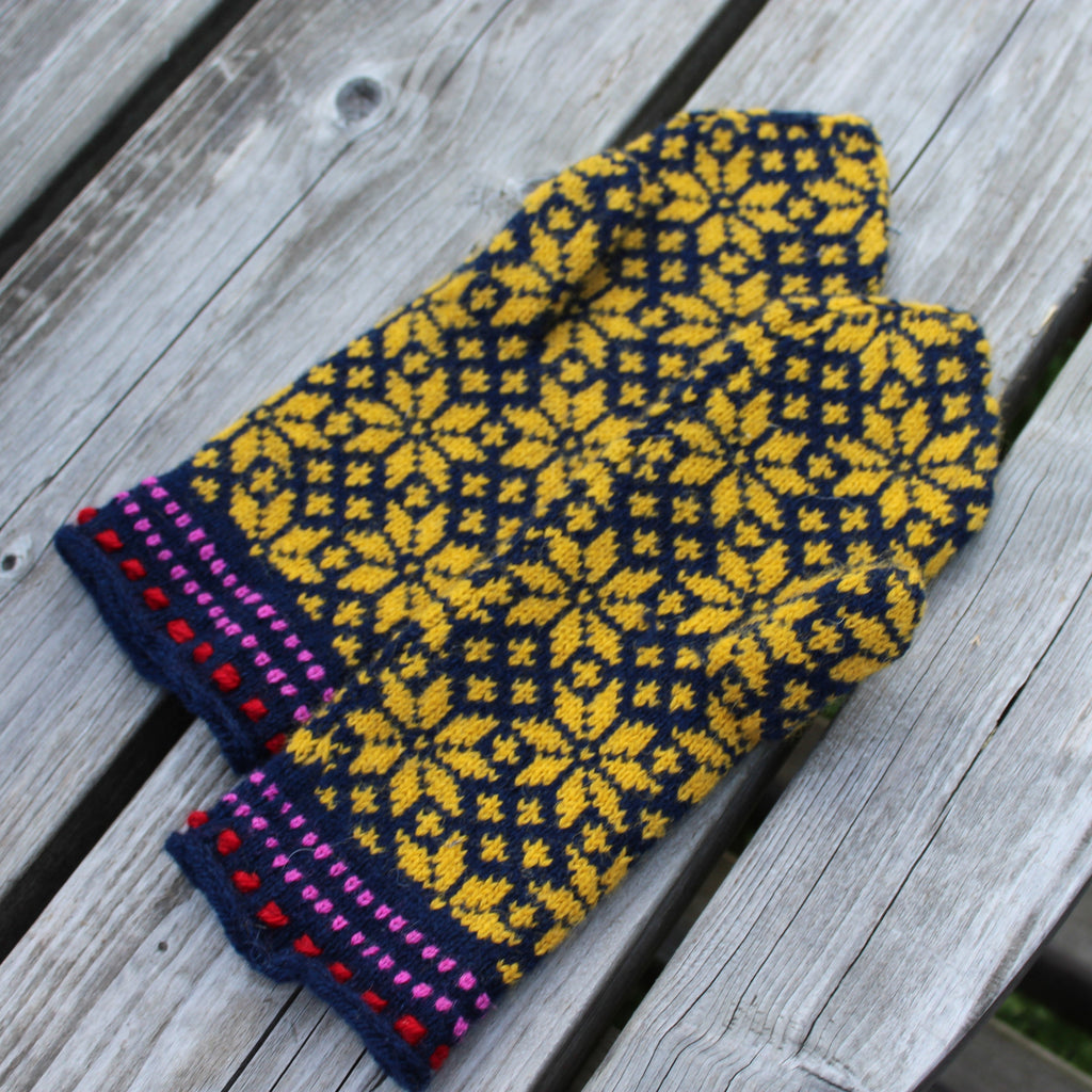 A pair of hand-knit pet mittens with a yellow and navy blue geometric pattern and colorful striped trim, displayed on a weathered gray wooden surface.
