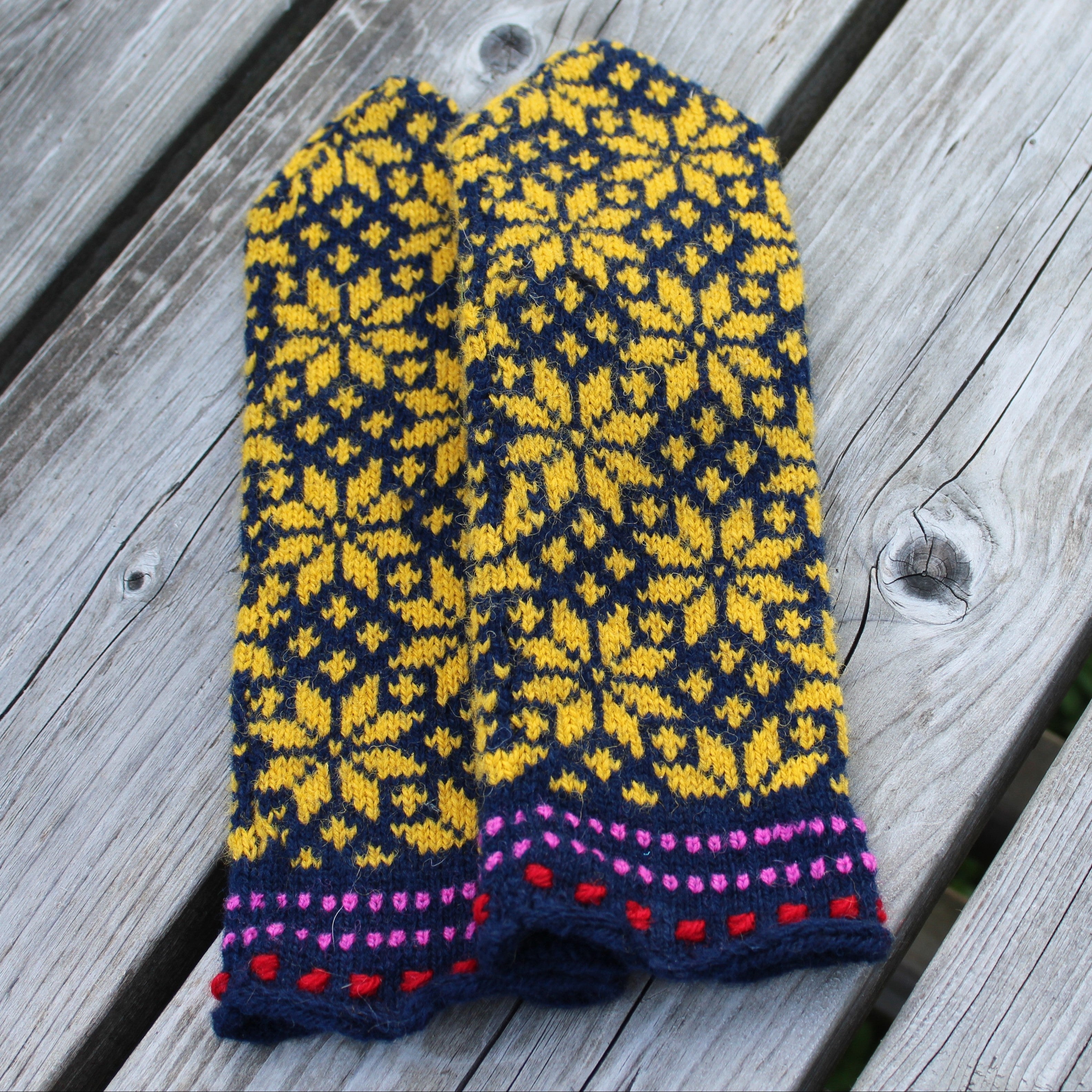 A pair of hand-knit winter mittens with a yellow and navy blue Fair Isle snowflake pattern and a pink and red dotted cuff, laid flat on weathered gray wooden planks.