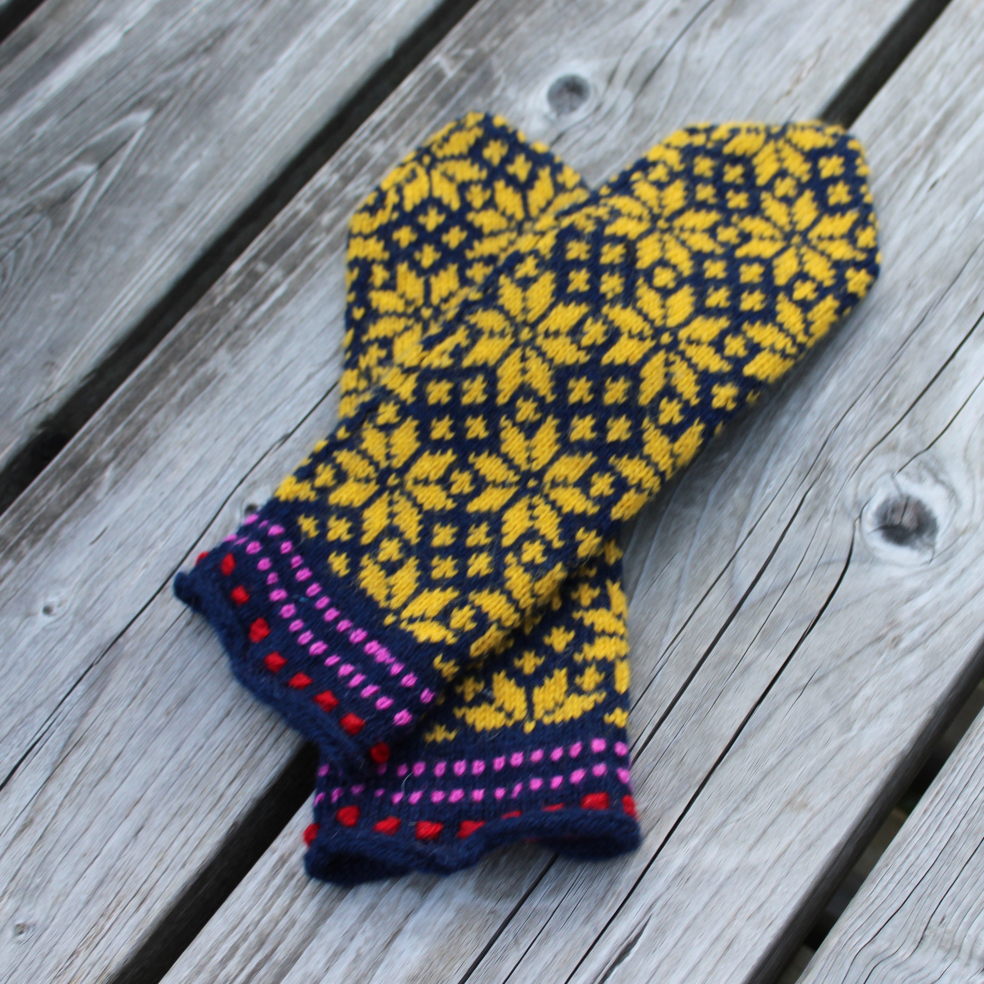 A pair of hand-knit mittens with a yellow and dark navy geometric pattern and a purple and pink striped cuff, displayed on a gray wooden plank surface.