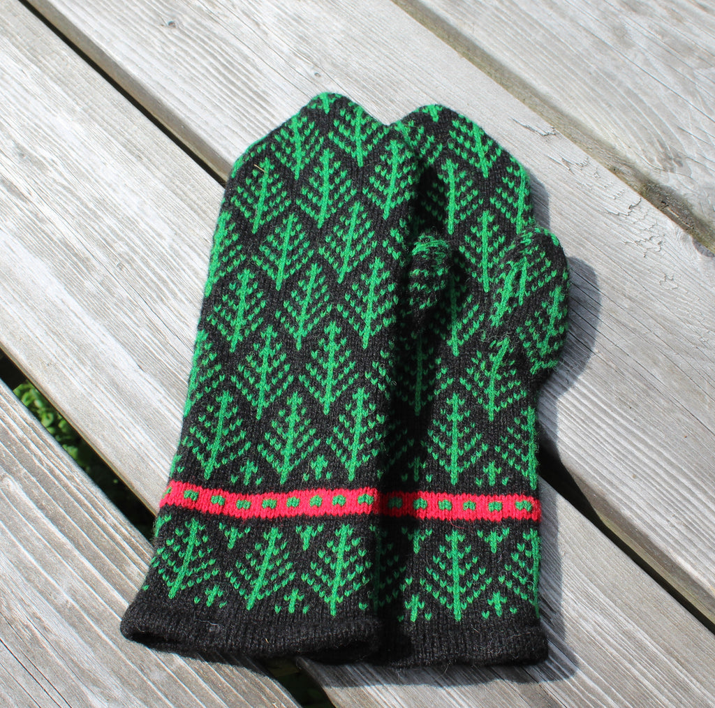 A pair of hand-knit winter mittens with a green and black tree pattern and a red stripe detail, displayed on a weathered gray wooden deck.