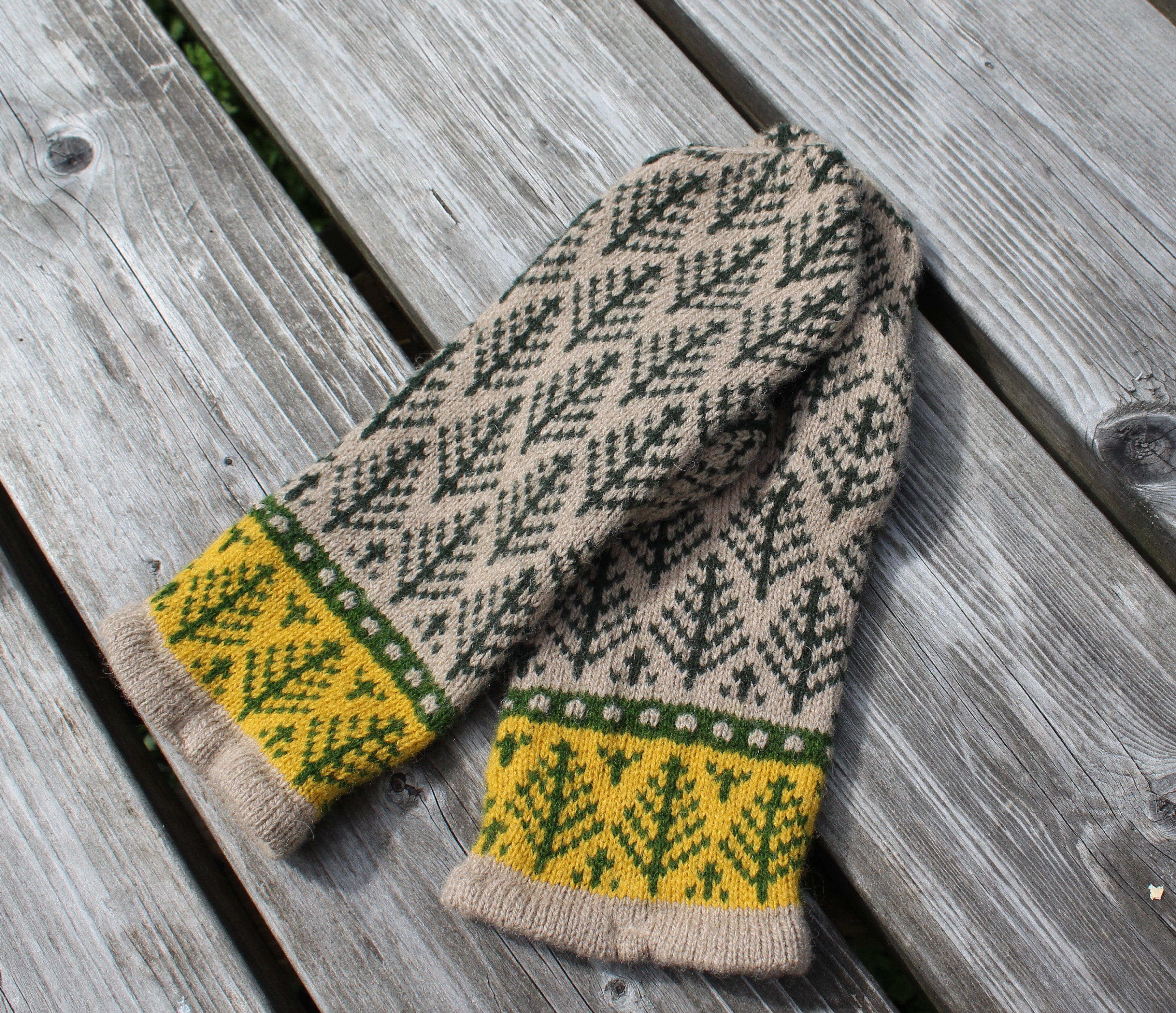 A hand-knit scarf with a beige and dark green tree pattern and yellow cuffs, laid flat on weathered gray wooden planks.