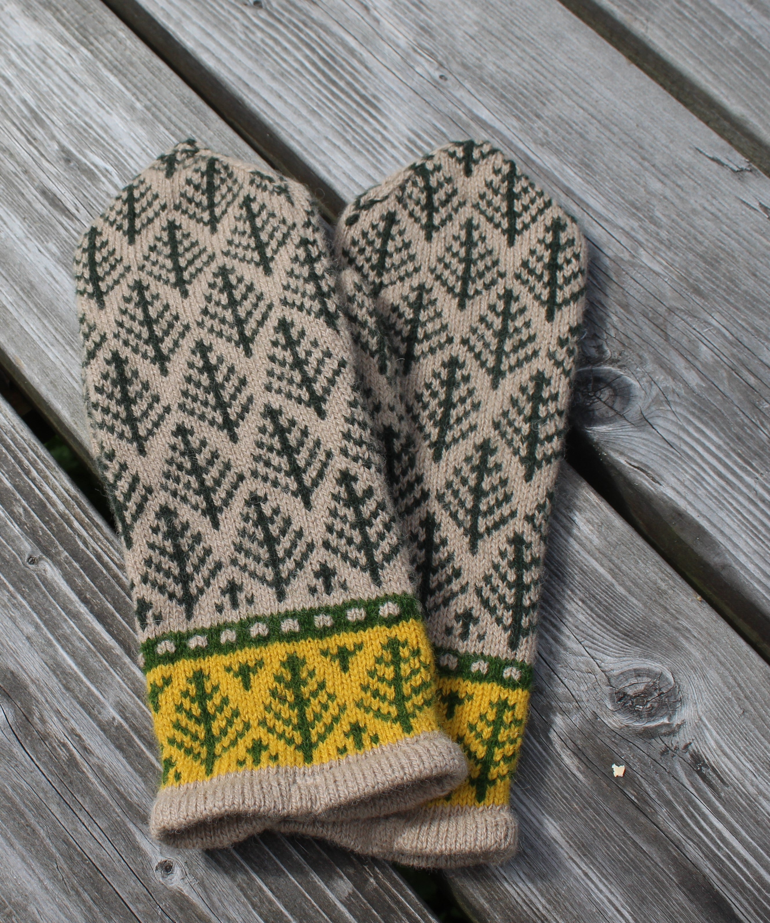 A pair of knit mittens with a dark green tree pattern on a beige background, featuring a yellow and green patterned cuff, laid flat on weathered gray wooden planks.