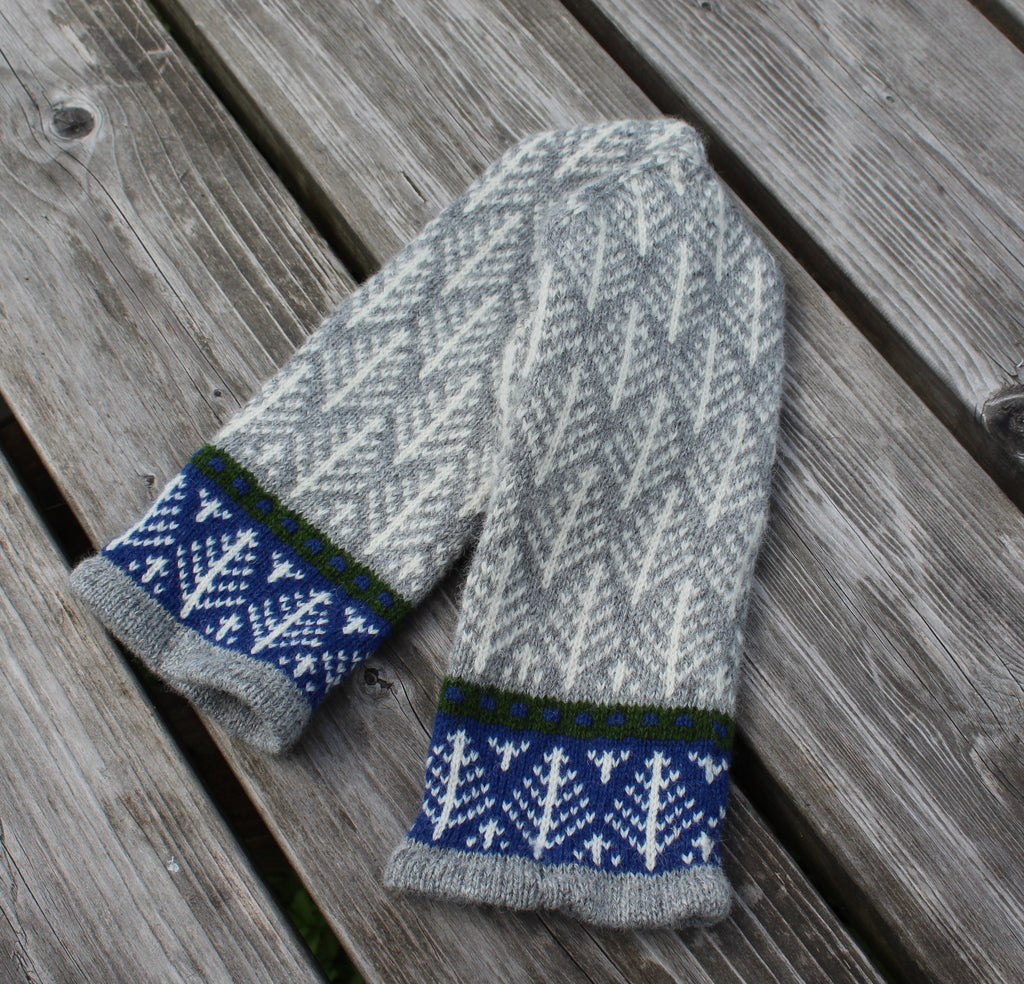 A pair of hand-knit winter mittens in gray and white with blue and green patterned cuffs, laid flat on weathered wooden planks.