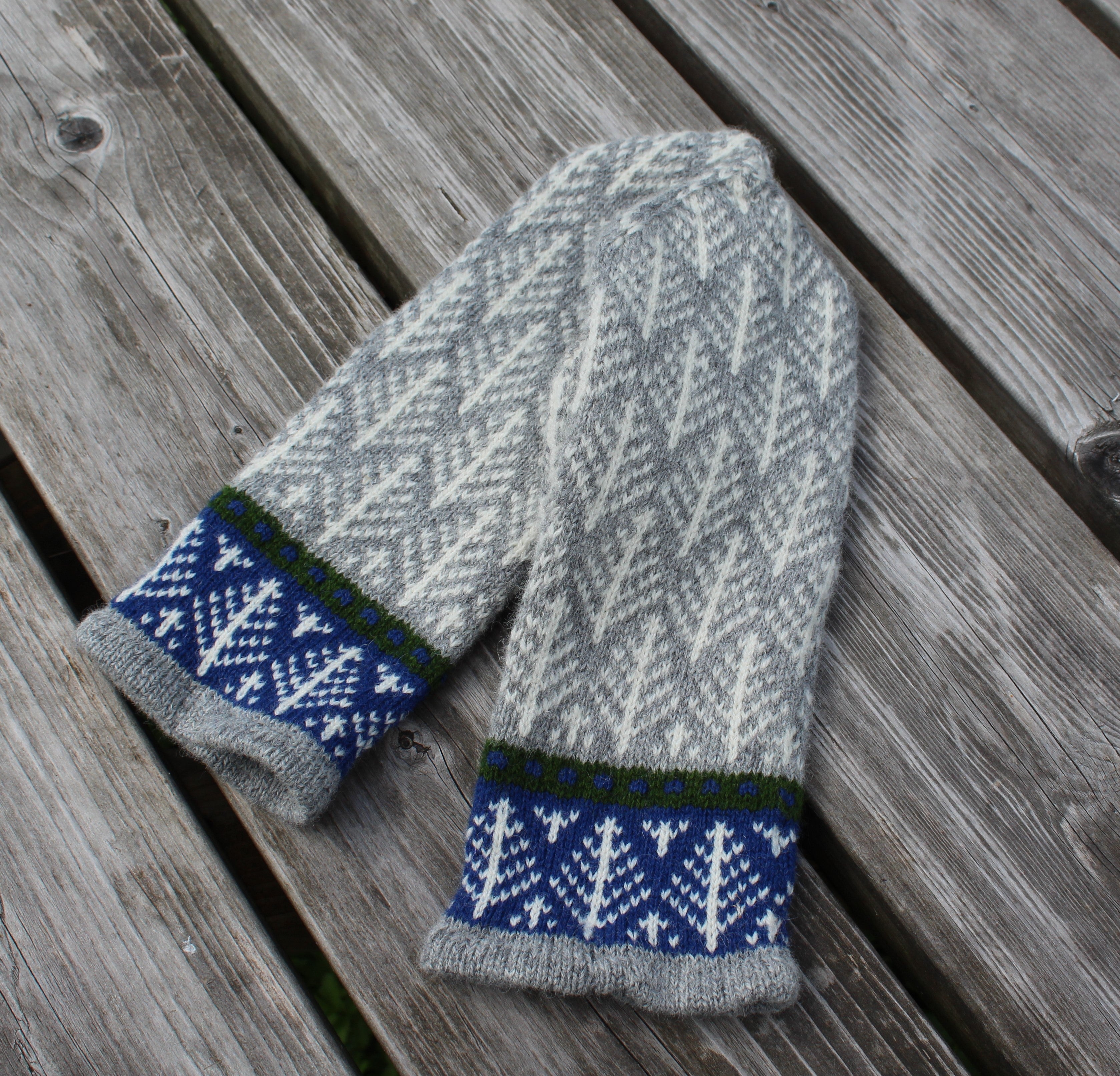 A pair of hand-knit winter mittens in gray and white with blue and green patterned cuffs, laid flat on weathered wooden planks.
