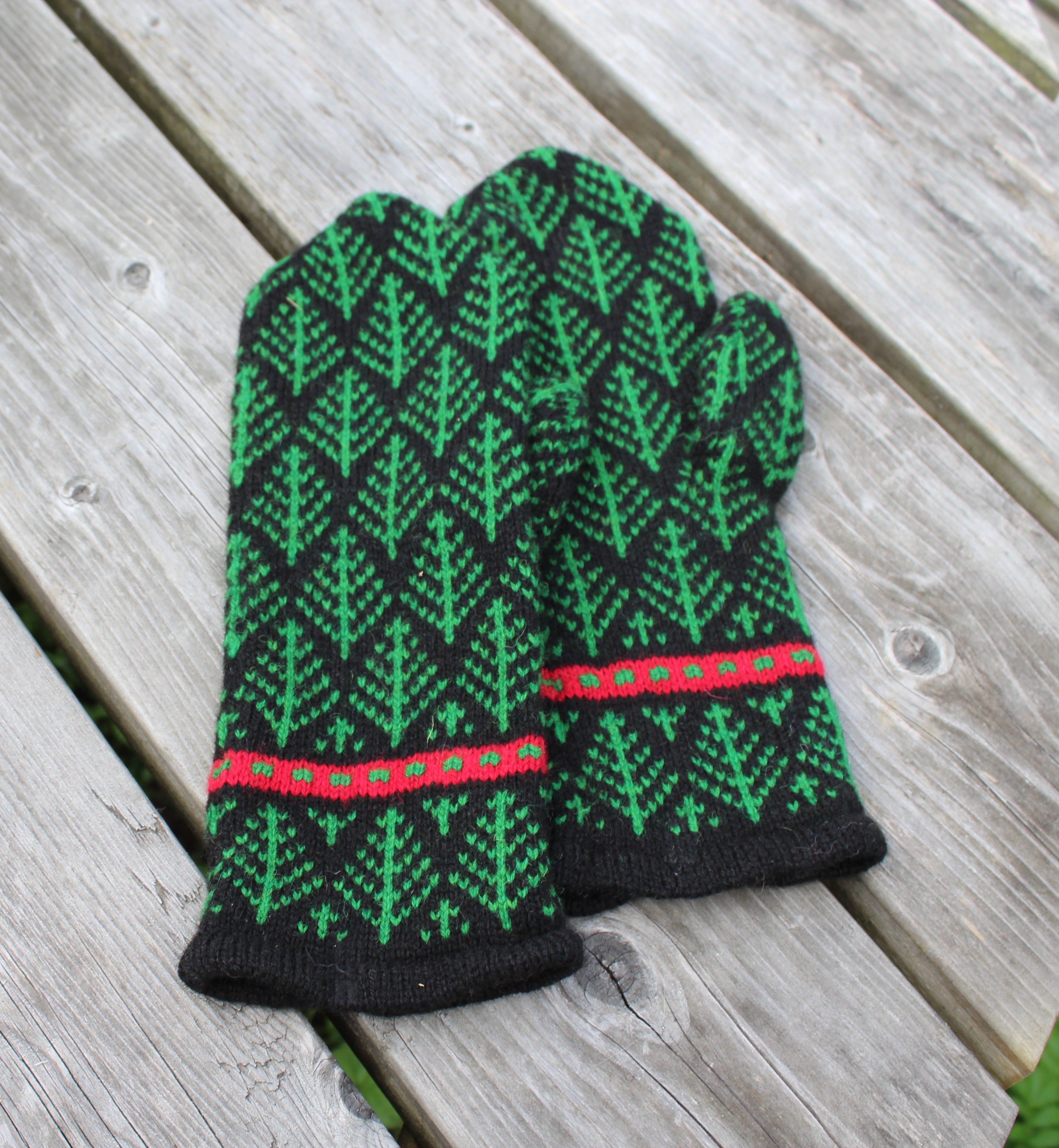 A pair of knit winter gloves with a green and black tree pattern and red accents, laid flat on a weathered gray wooden plank surface.