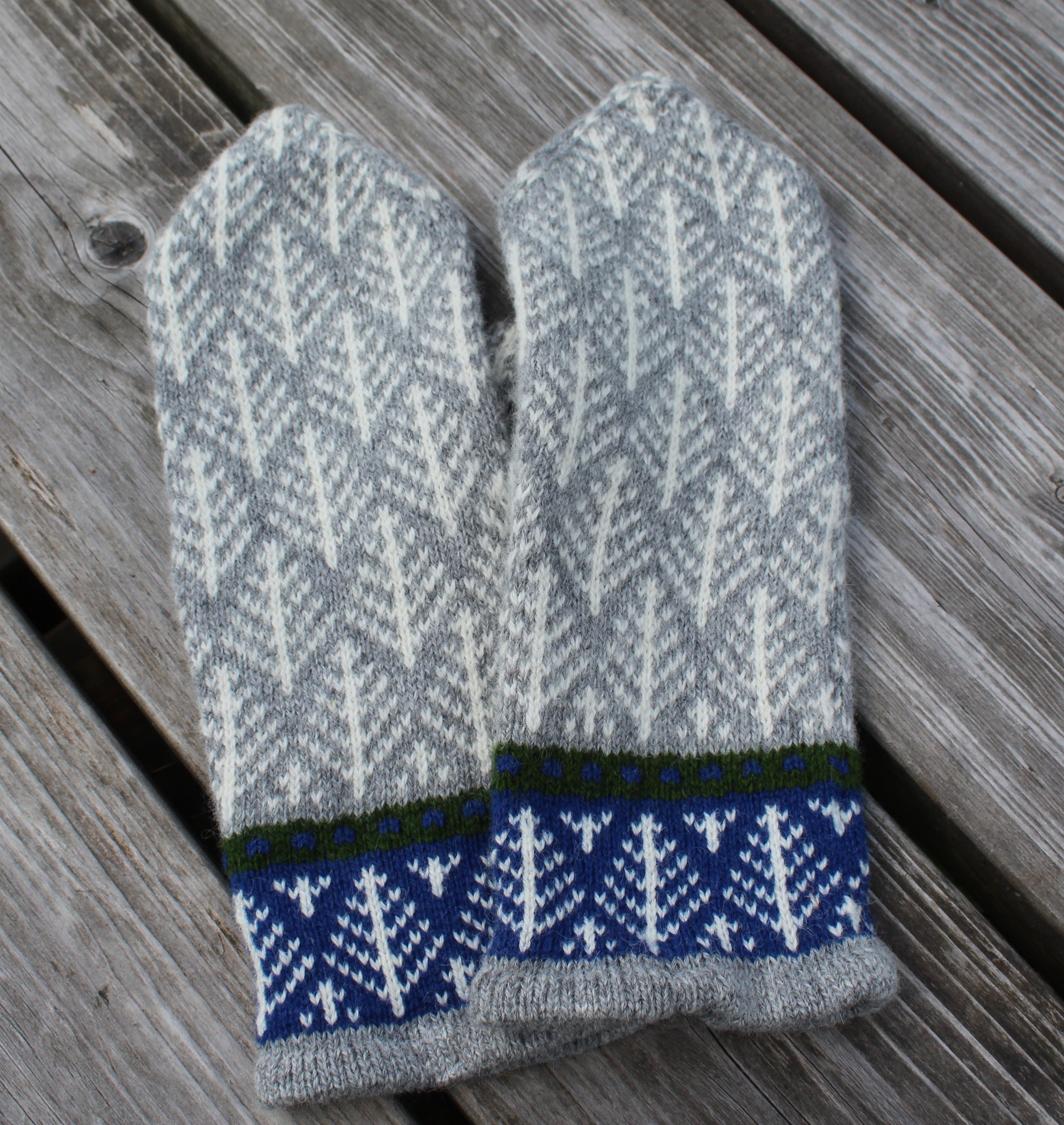 A pair of knit mittens with a gray and white geometric pattern and a blue patterned cuff, laid flat on weathered wooden planks.