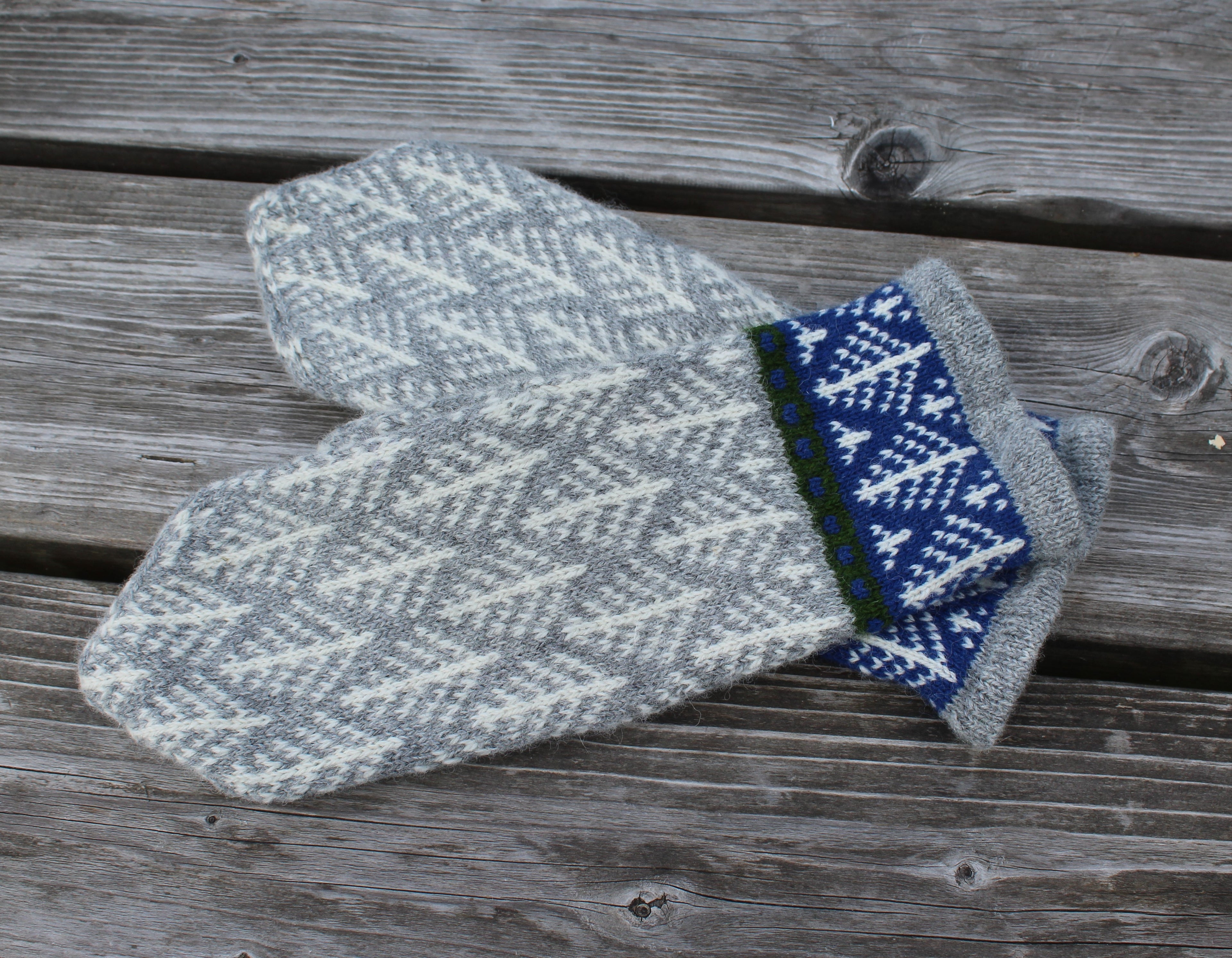 A pair of knit winter mittens with a gray and white geometric pattern, featuring a folded cuff in blue, white, and green, displayed on a weathered wooden plank surface.