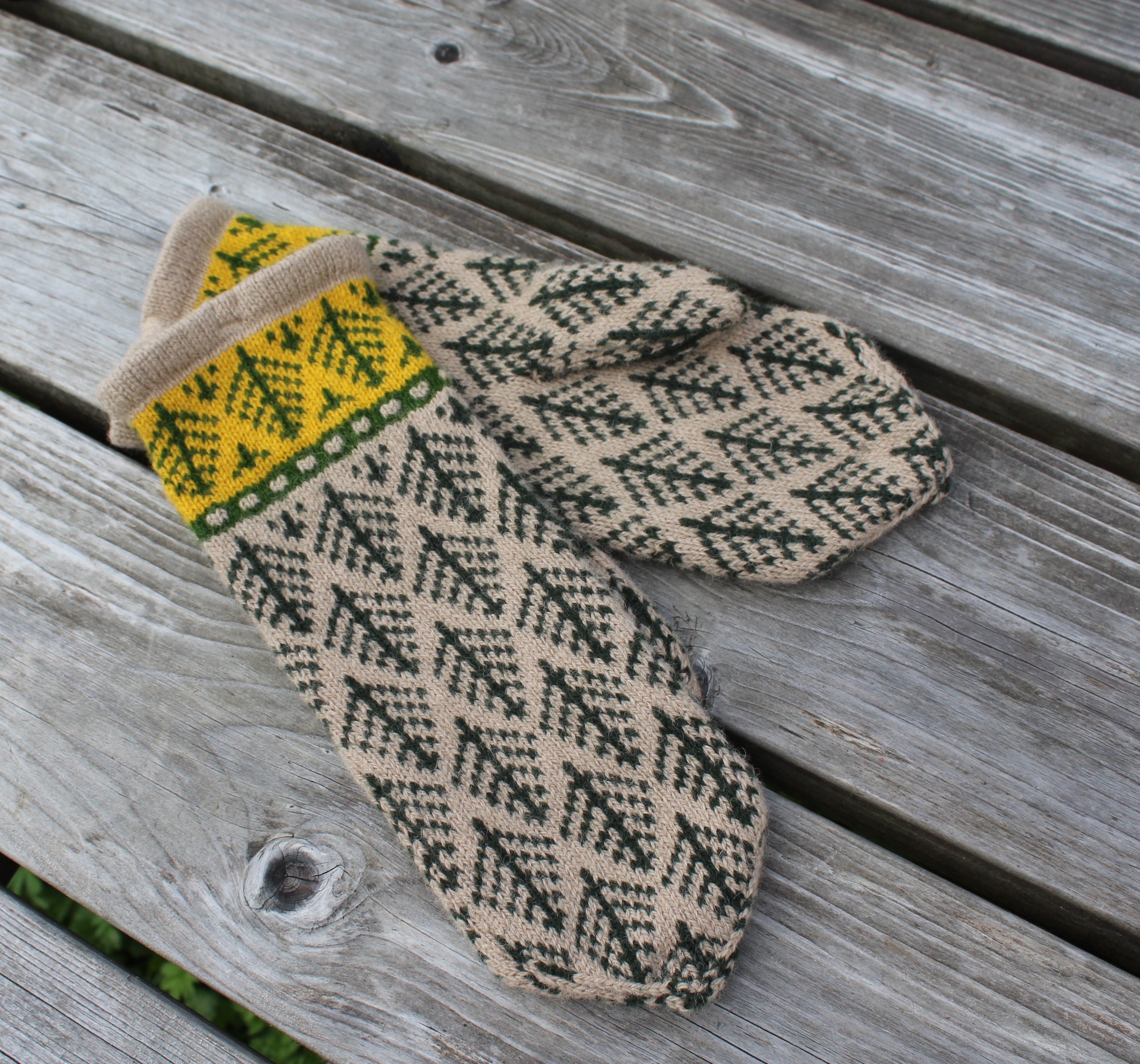 A pair of knit winter mittens with a beige base, dark green geometric tree pattern, and a yellow and green patterned cuff, displayed on a weathered gray wooden deck.