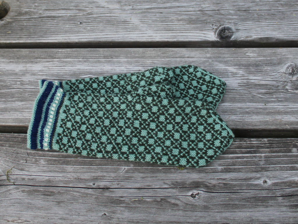 A single knit mitten in a green and black pattern with a striped cuff, laid flat on weathered gray wooden planks.