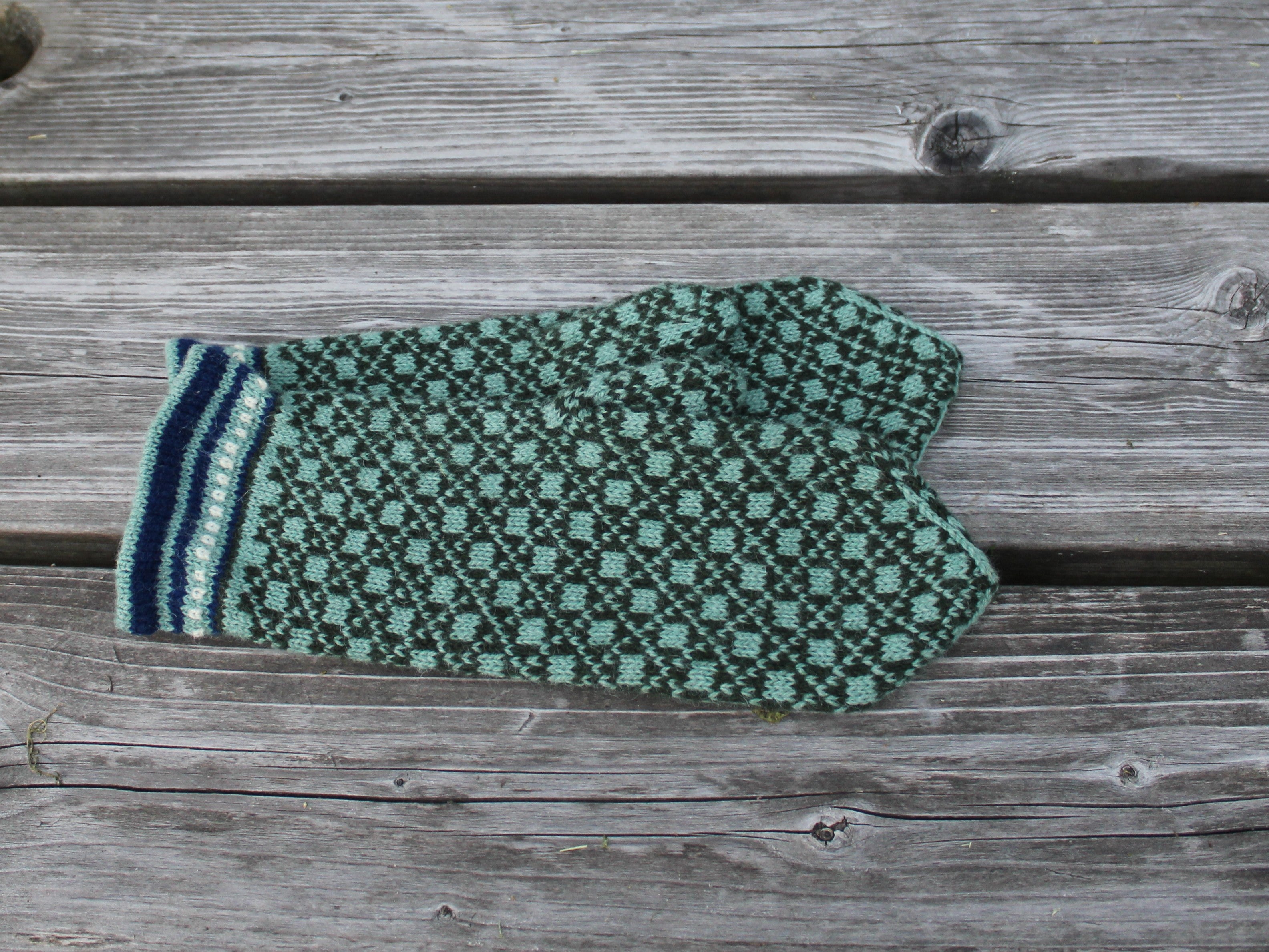 A single knit mitten in a green and black pattern with a striped cuff, laid flat on weathered gray wooden planks.