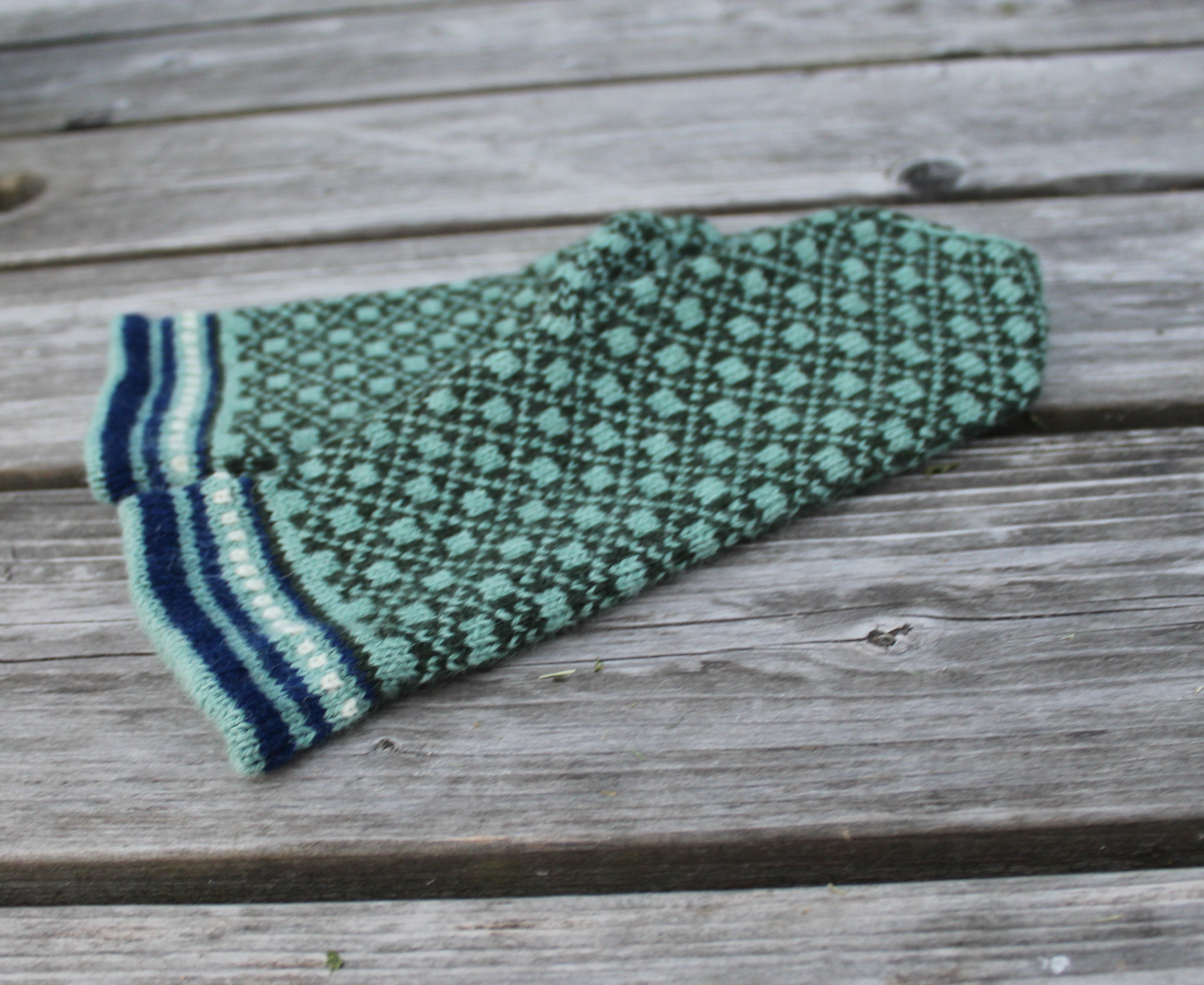 A green and black patterned knit sock with navy and cream striped cuffs, laid flat on weathered wooden planks.