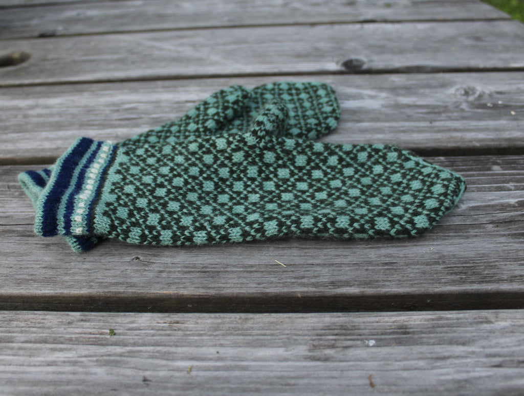 A single knit mitten in green and navy with a geometric pattern and striped cuff, resting on weathered gray wooden planks.