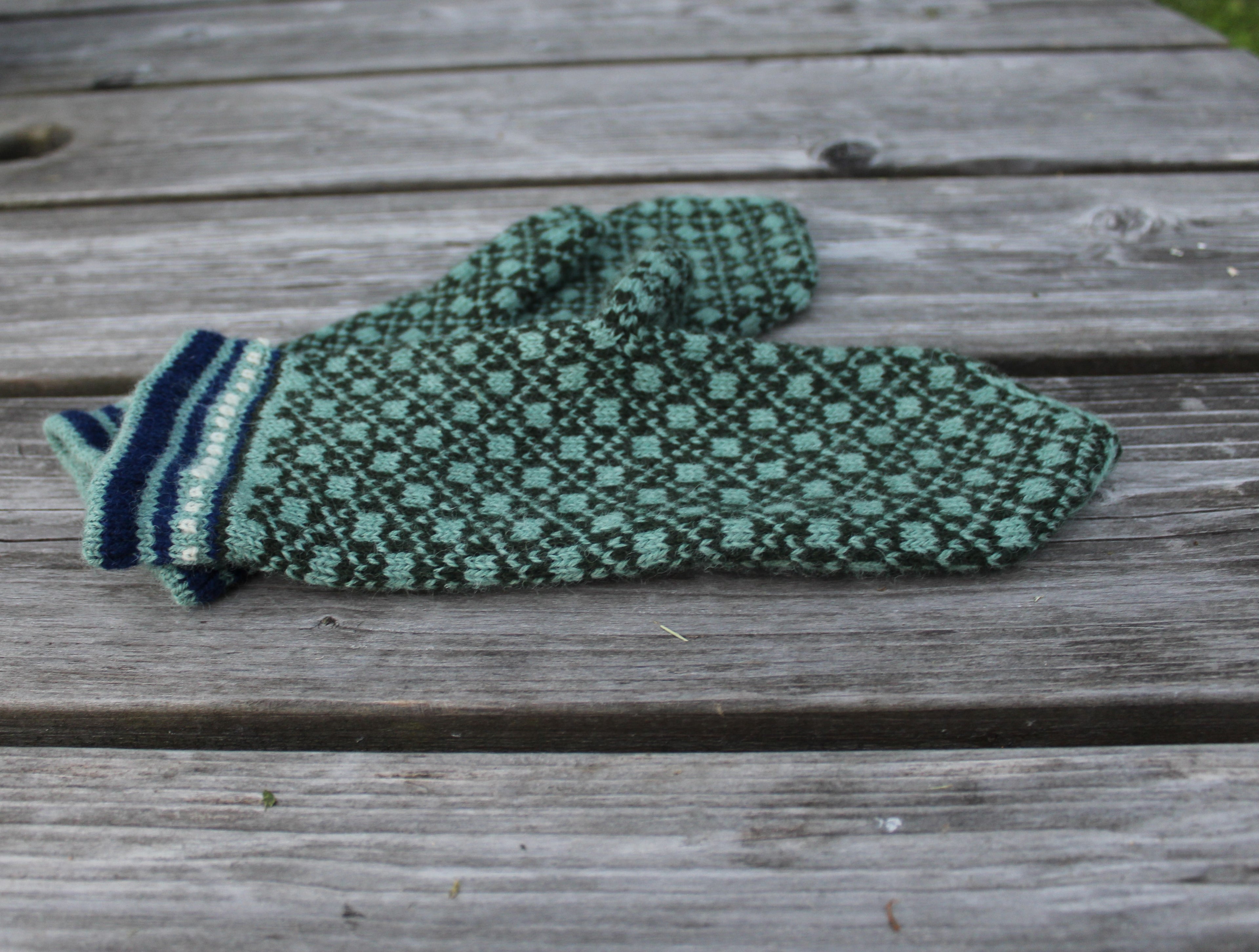 A single knit mitten in green and navy with a geometric pattern and striped cuff, resting on weathered gray wooden planks.