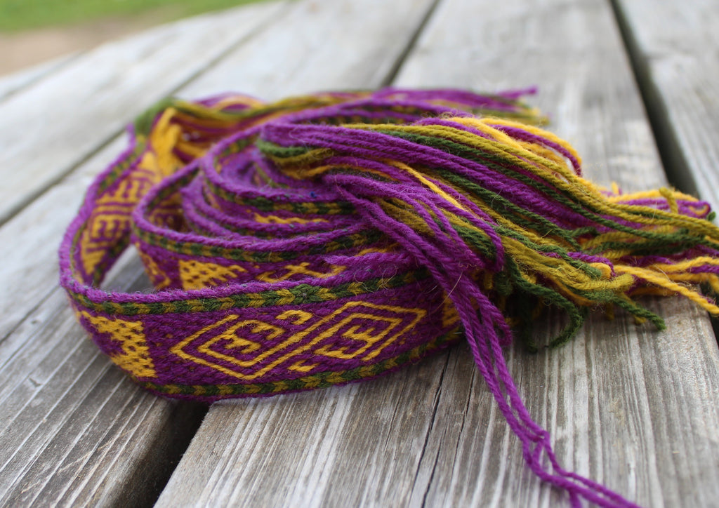 Card weaving latvian belt featuring purple, yellow, and green geometric patterns, resting on weathered gray wooden planks.