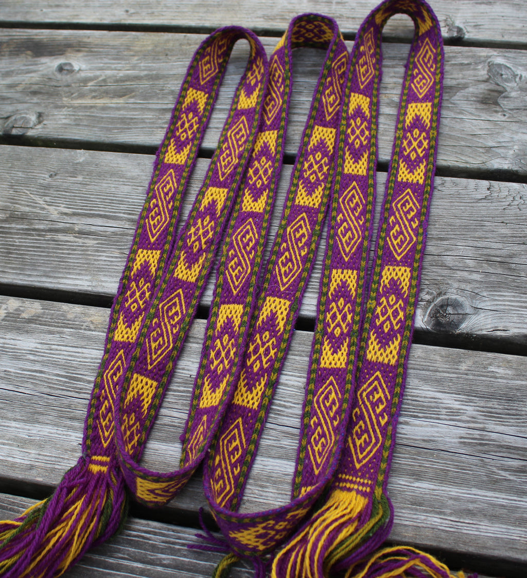 A handmade woven latvian belt with purple, yellow, and green geometric patterns and tassels, laid out on a weathered gray wooden plank surface.