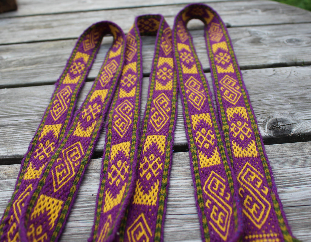 A close-up of a woven textile strap or belt laid flat on a weathered gray wooden surface. The fabric has a repeating geometric pattern in purple and yellow with thin green edging.