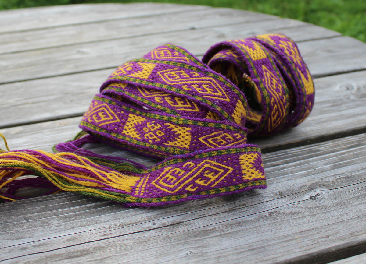 A woven Latvian  with purple, yellow, and green geometric patterns, resting on a weathered gray wooden table with green grass in the background.ool belt