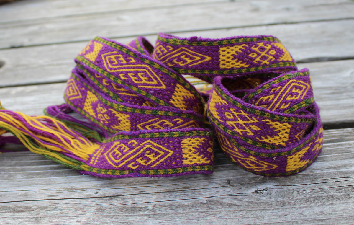 A coiled multicolor woven textile with geometric patterns in purple, yellow, and green, resting on a weathered gray wooden surface.