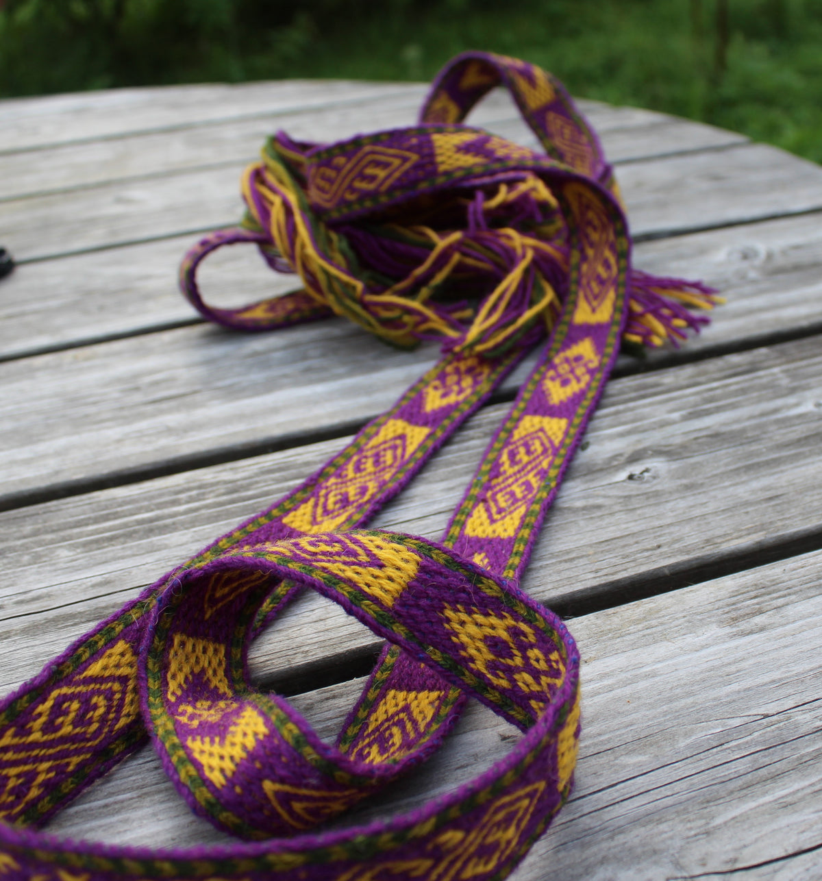 A woven textile strap with a purple and yellow geometric pattern, draped on a weathered wooden table with a blurred green outdoor background.