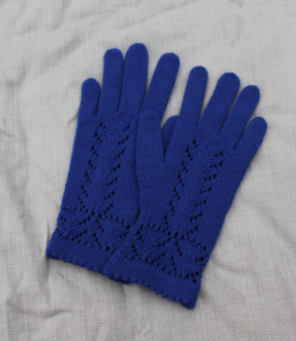 A pair of royal blue hand-knit gloves with an intricate openwork leaf pattern on the back of the hands and a ribbed cuff, laid flat on a textured light gray fabric background.