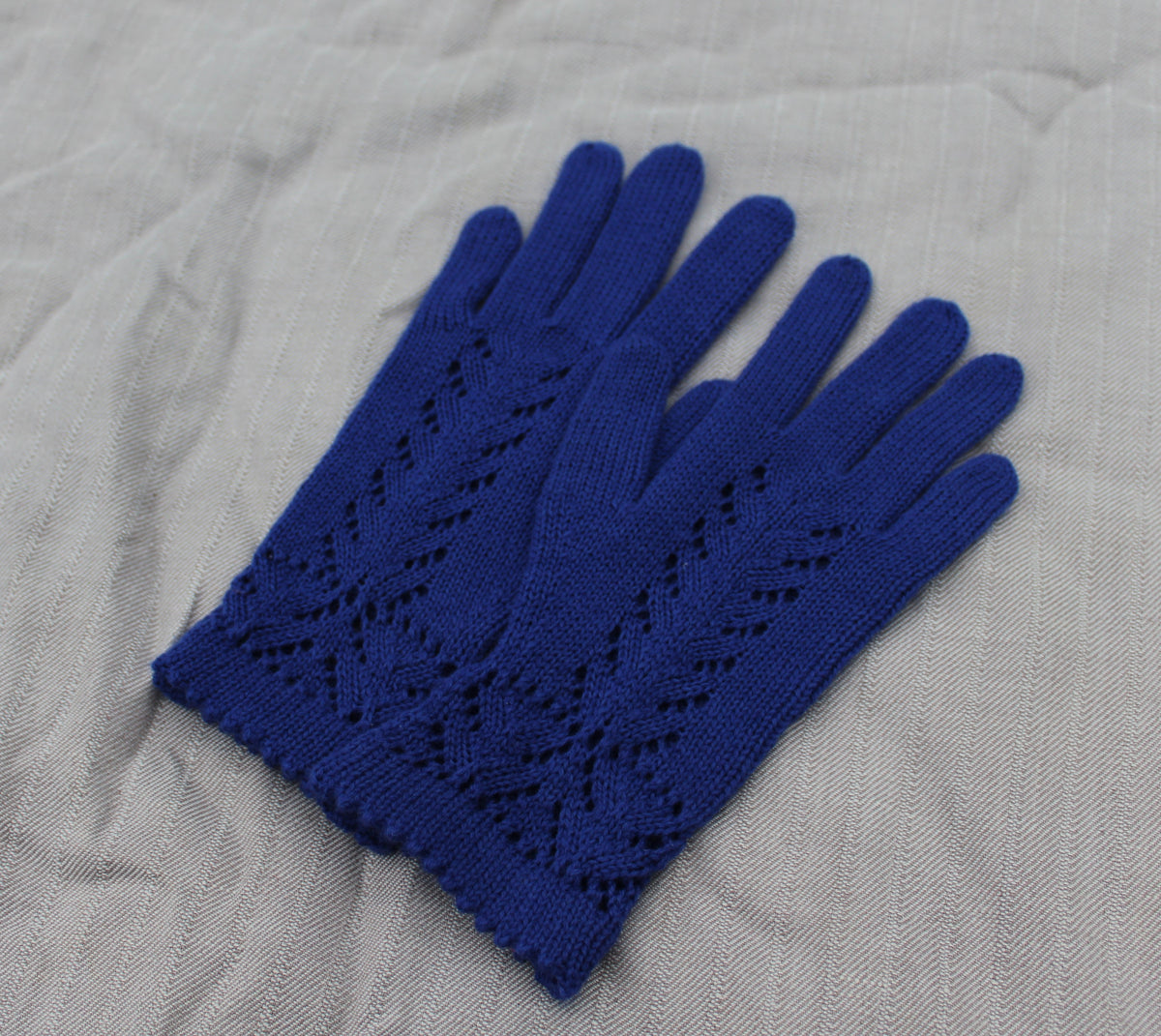 A pair of royal blue knit gloves with an openwork lace pattern on the backs, laid flat on a textured light gray fabric background.