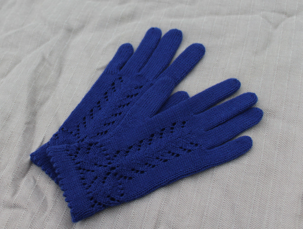 A pair of royal blue knit gloves with an openwork leaf pattern on the backs, laid flat on a textured light gray fabric background.