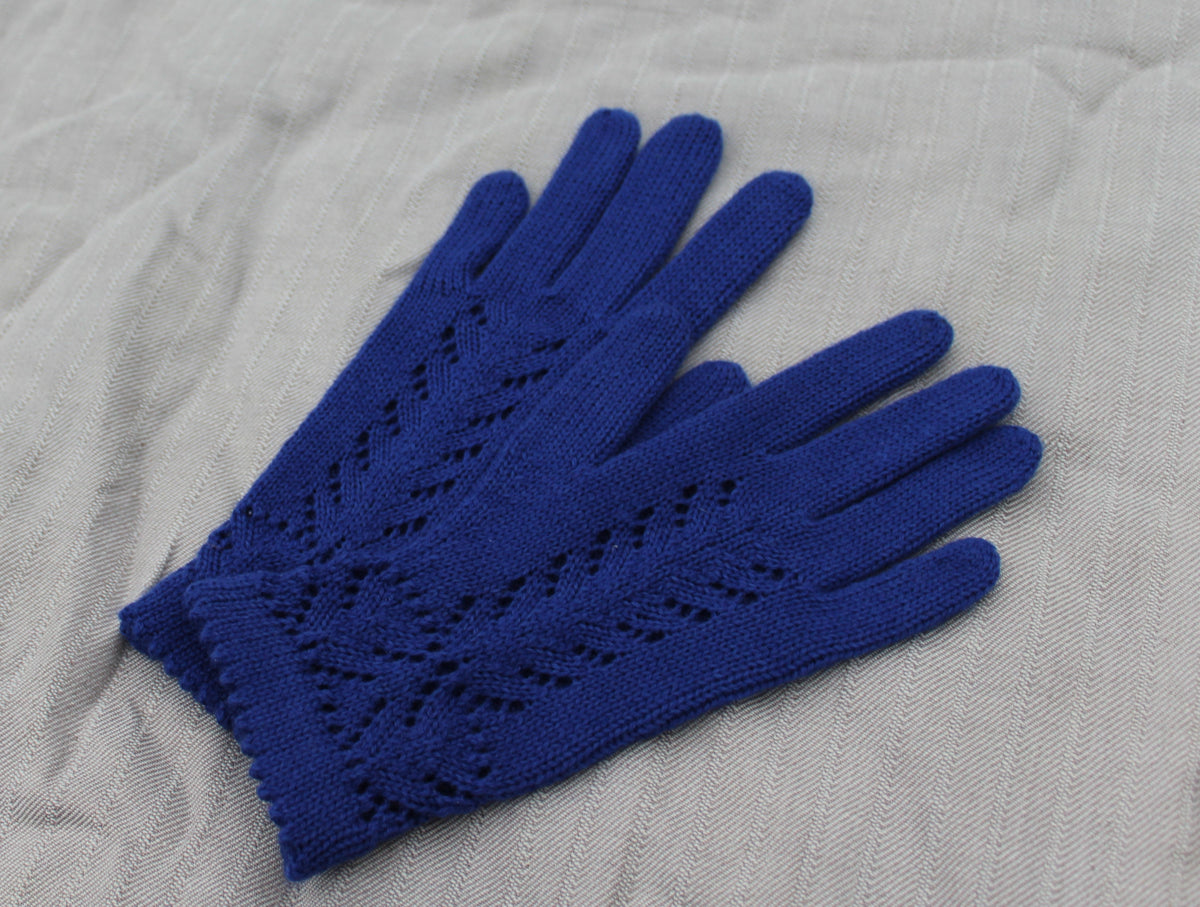 A pair of royal blue knit gloves with an openwork leaf pattern on the backs, laid flat on a textured light gray fabric background.