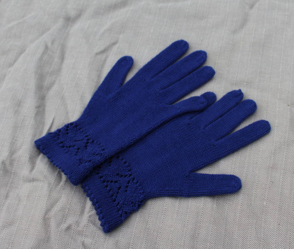 A pair of royal blue hand-knit gloves with a decorative cable-knit cuff, laid flat on a textured light gray fabric background.