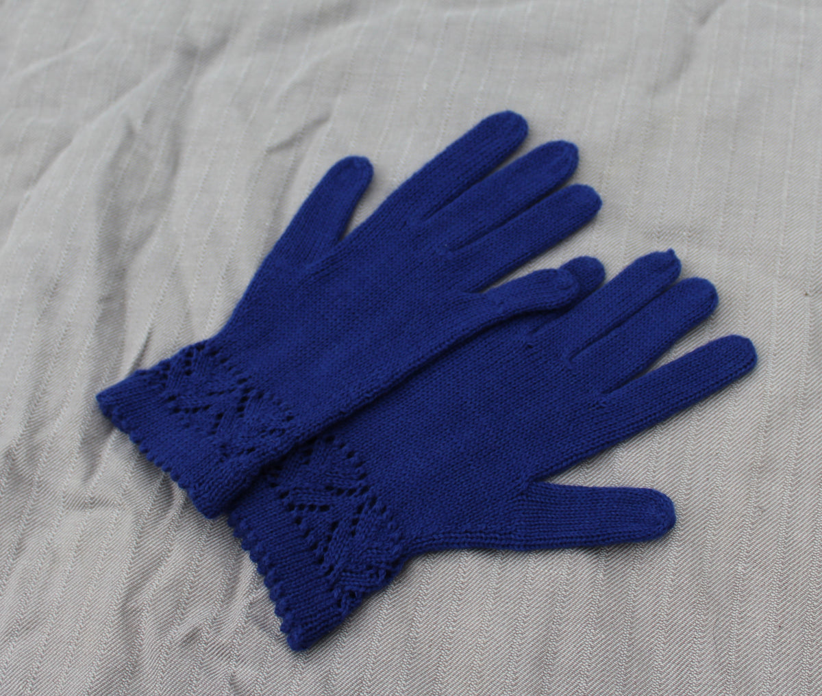 A pair of royal blue hand-knit gloves with a decorative cable-knit cuff, laid flat on a textured light gray fabric background.