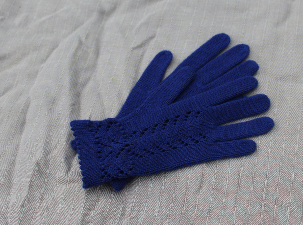 A pair of royal blue knit gloves with a textured, openwork chevron pattern on the back of the hand, laid flat on a light gray textured fabric background.