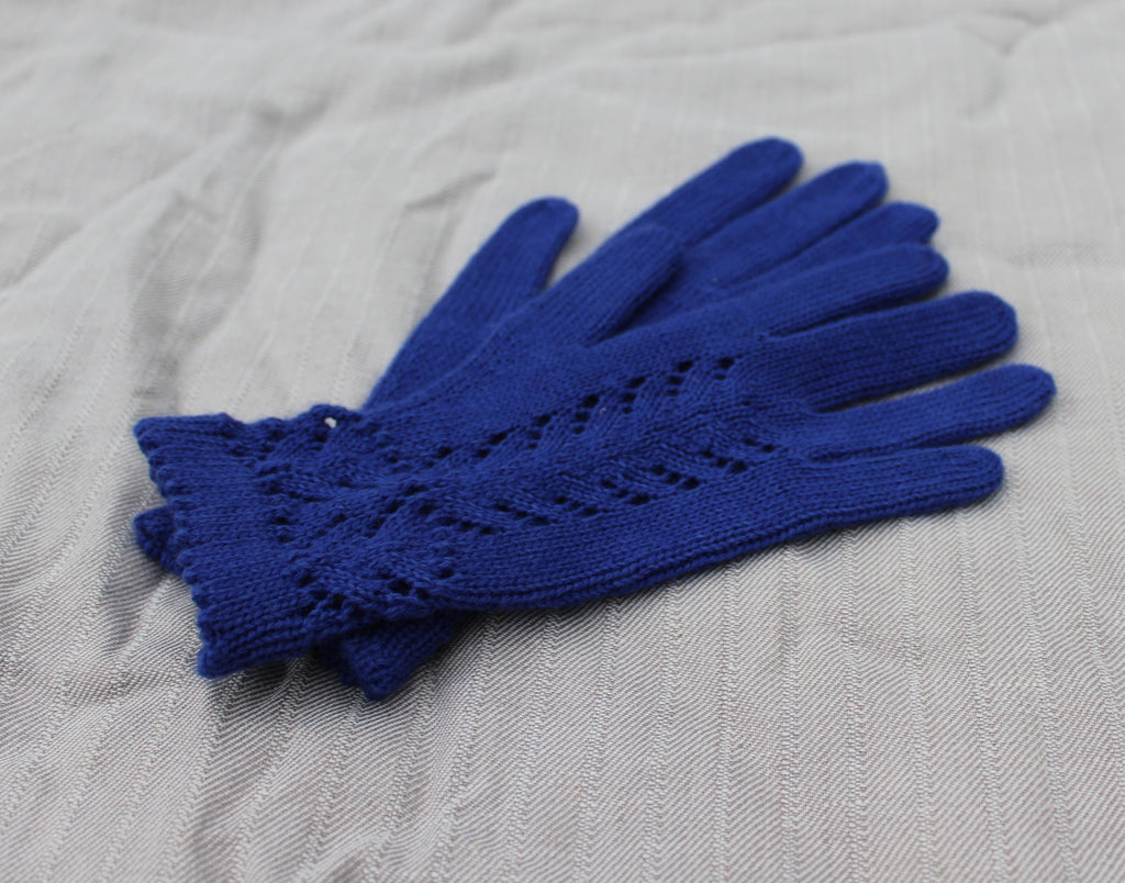 A pair of royal blue knit gloves with a lace-like openwork pattern at the cuff, laid flat on a textured light gray fabric background.