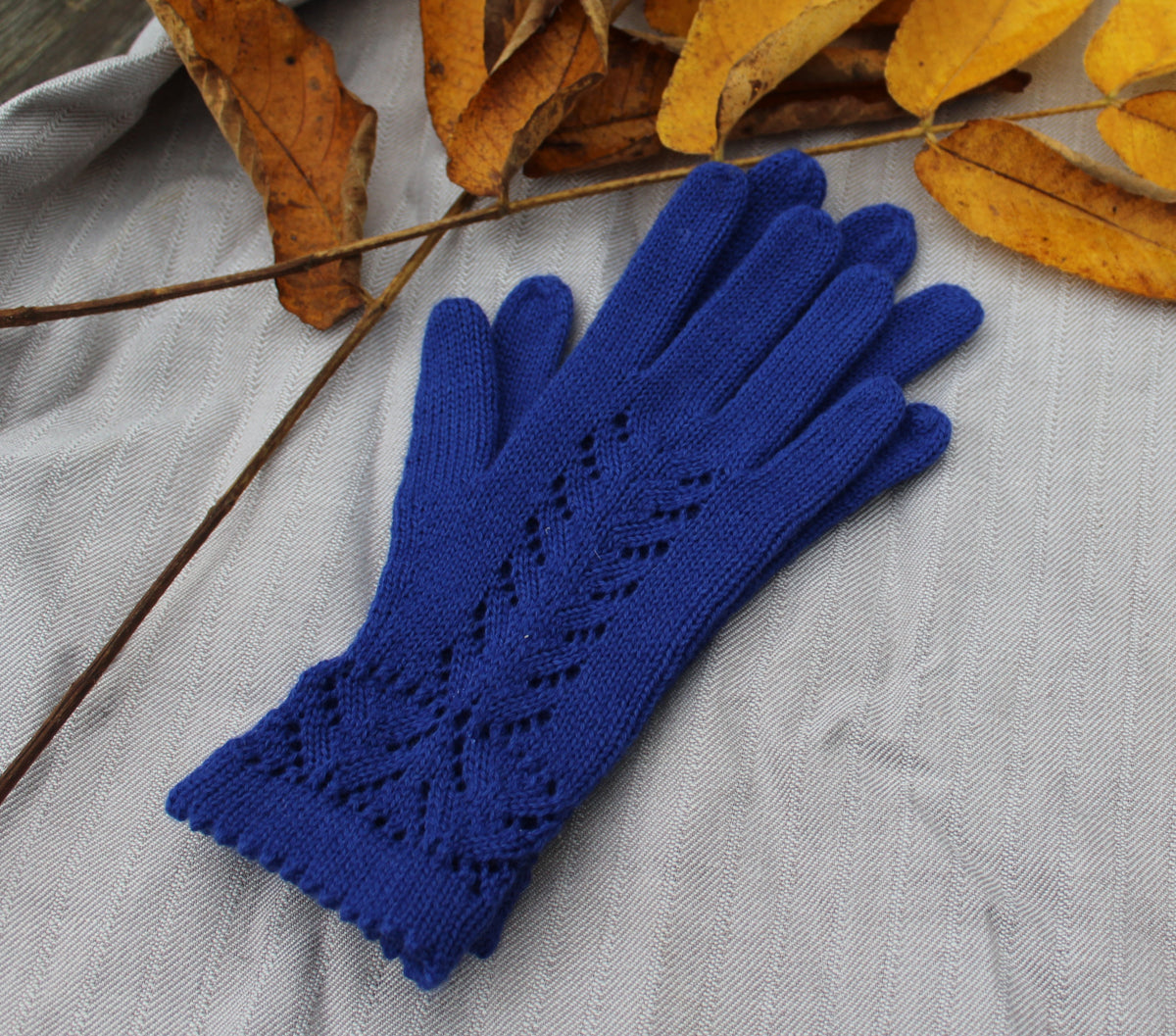 A single royal blue knit glove with a textured leaf pattern on the back of the hand, laid flat on a gray textured fabric background with scattered brown and yellow autumn leaves.